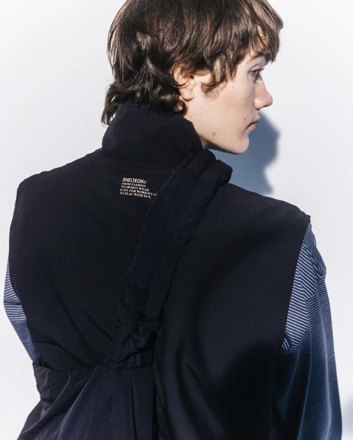 A young person with short, wavy brown hair and fair skin, wearing a black high-collar jacket with a small white label on the back, and a blue and white striped shirt underneath, standing against a plain white background, looking to the right in profile.