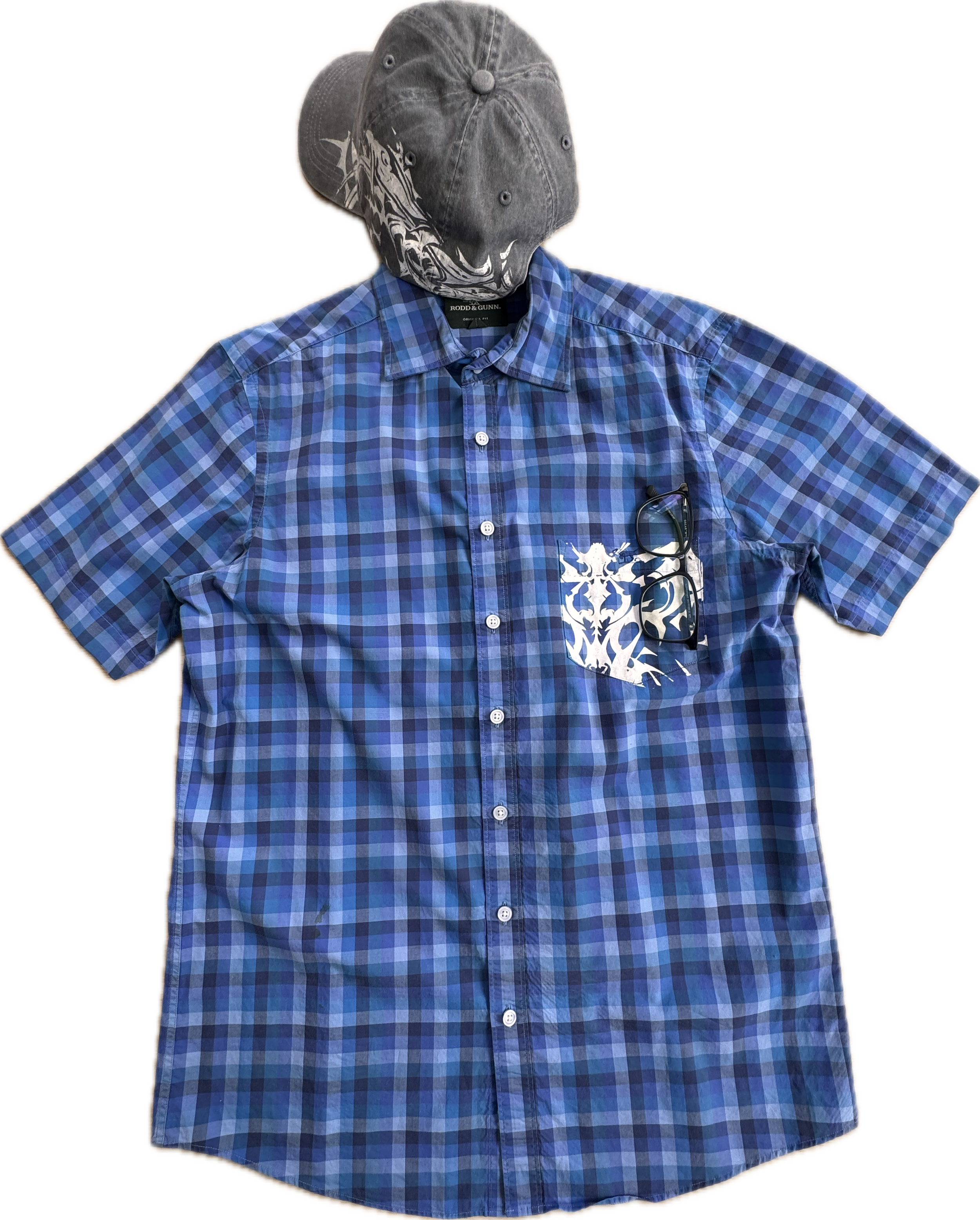 A short-sleeved, blue plaid button-up shirt with a white decorative pattern in the chest pocket, a pair of black-rimmed eyeglasses hanging from the collar, and a dark-colored baseball cap with a zebra graphic on it.