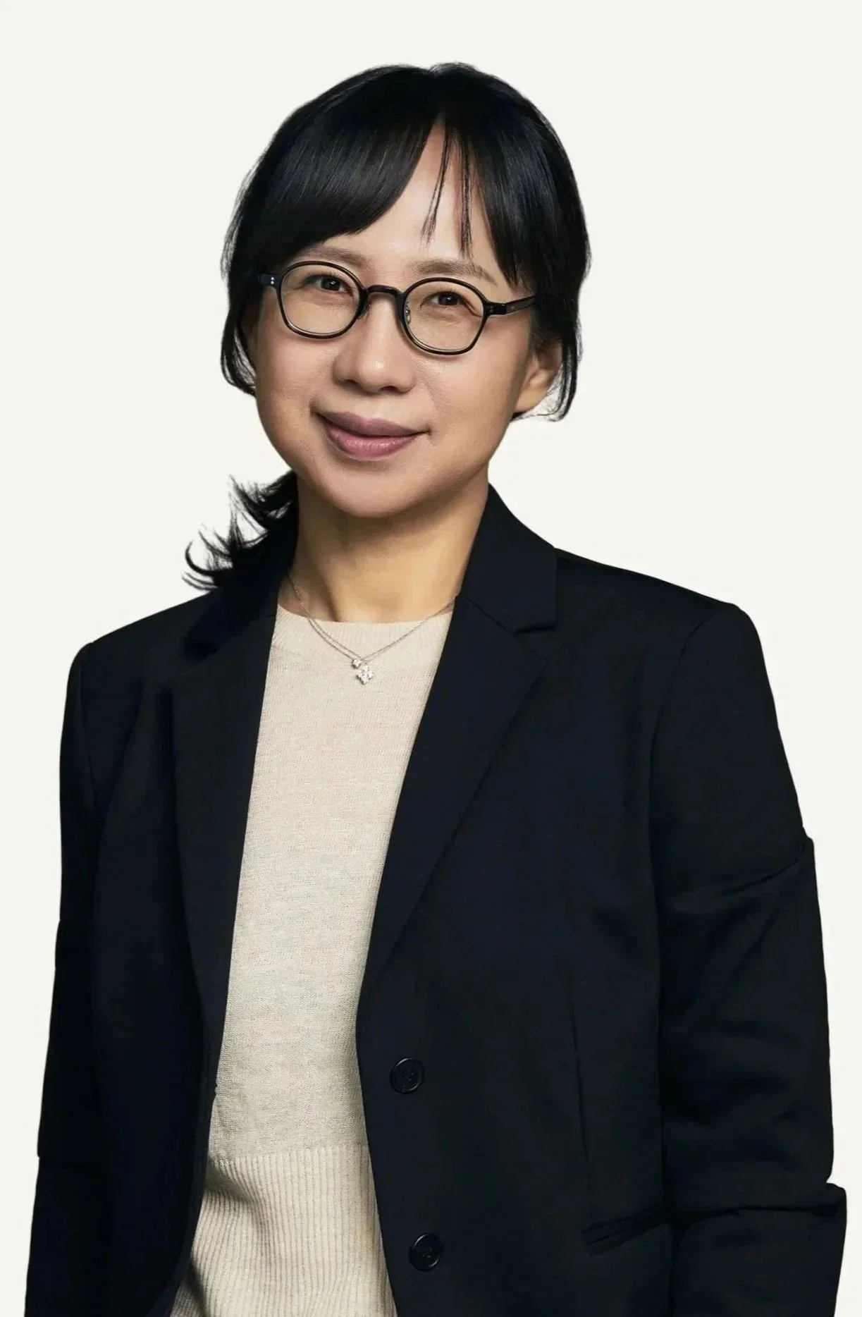 A woman with short black hair and glasses, wearing a black blazer and a beige top, smiling at the camera against a plain white background.
