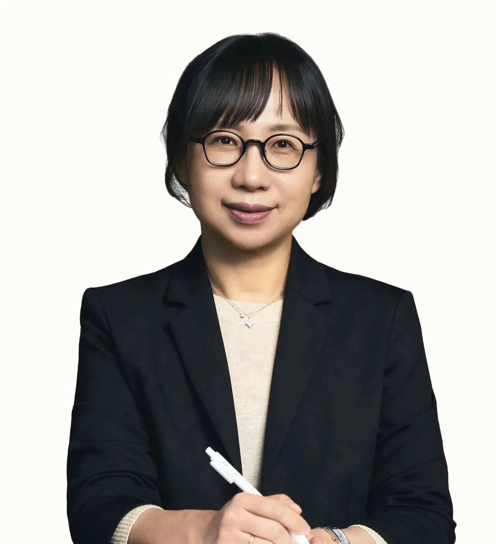 A woman with short black hair, wearing glasses, a black blazer, and a beige top, holding a white pen and smiling at the camera.