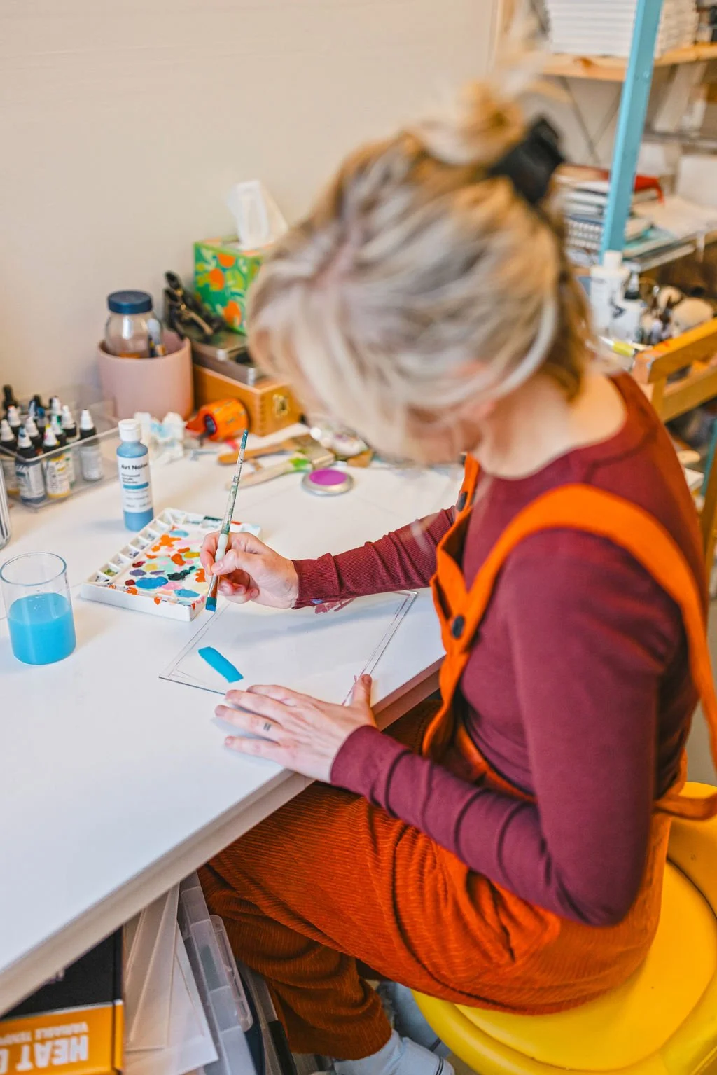 A woman sitting at a white table, painting on paper with watercolor paints, surrounded by art supplies and a glass of water.