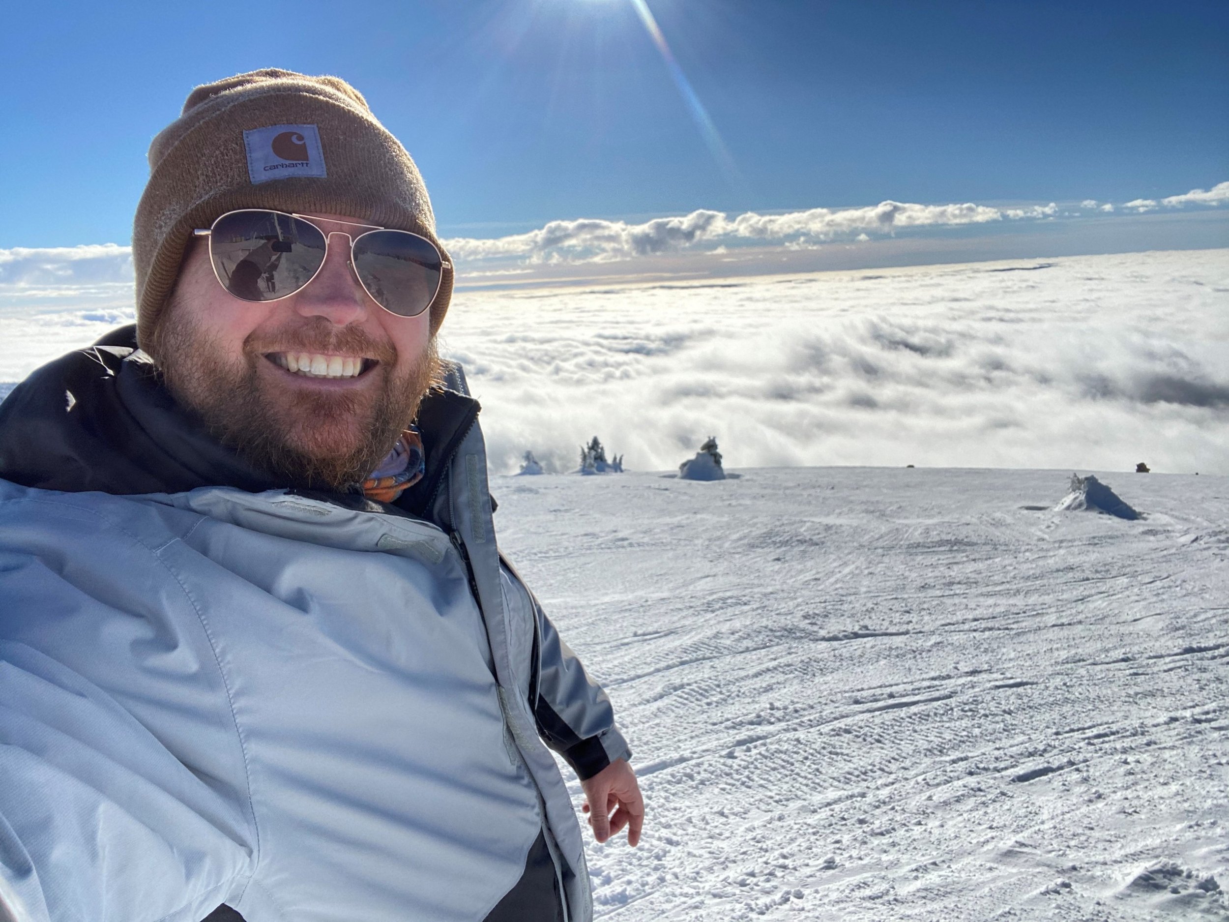 A man with a beard wearing sunglasses, a Carhartt beanie, and a winter jacket taking a selfie on a snowy mountain top with a cloudy sky and snow-covered trees in the background.