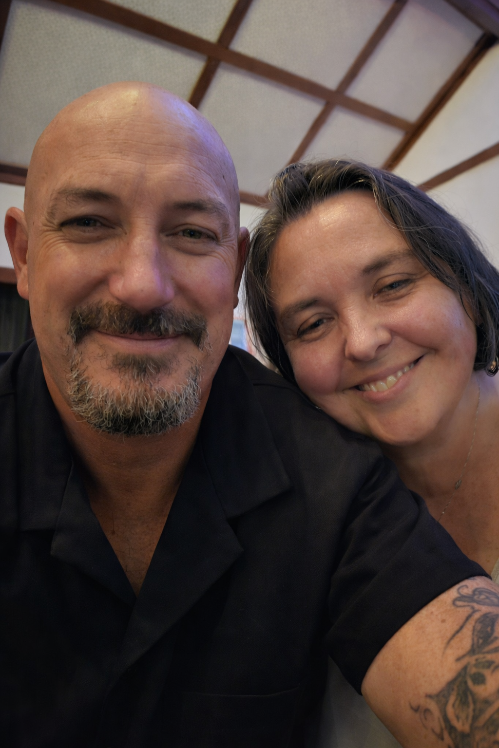 A close-up photo of a smiling man with a bald head, goatee, and mustache, and a woman with dark hair, both appearing happy and close together in an indoor setting.