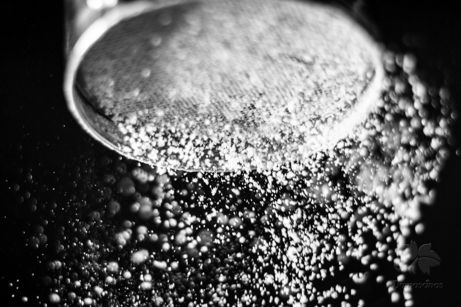 Close-up black and white photo of flour being poured from a strainer on dough and the flour is scattered in the air.