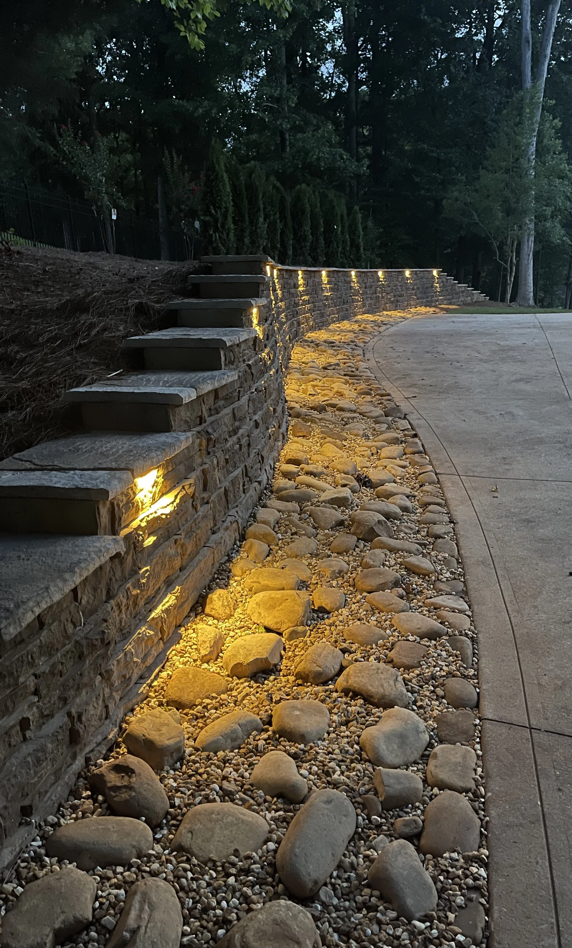 Curved stone retaining wall with integrated ground lighting, small rocks and pebbles along the base, surrounding a concrete walk and a wooded area in the background.
