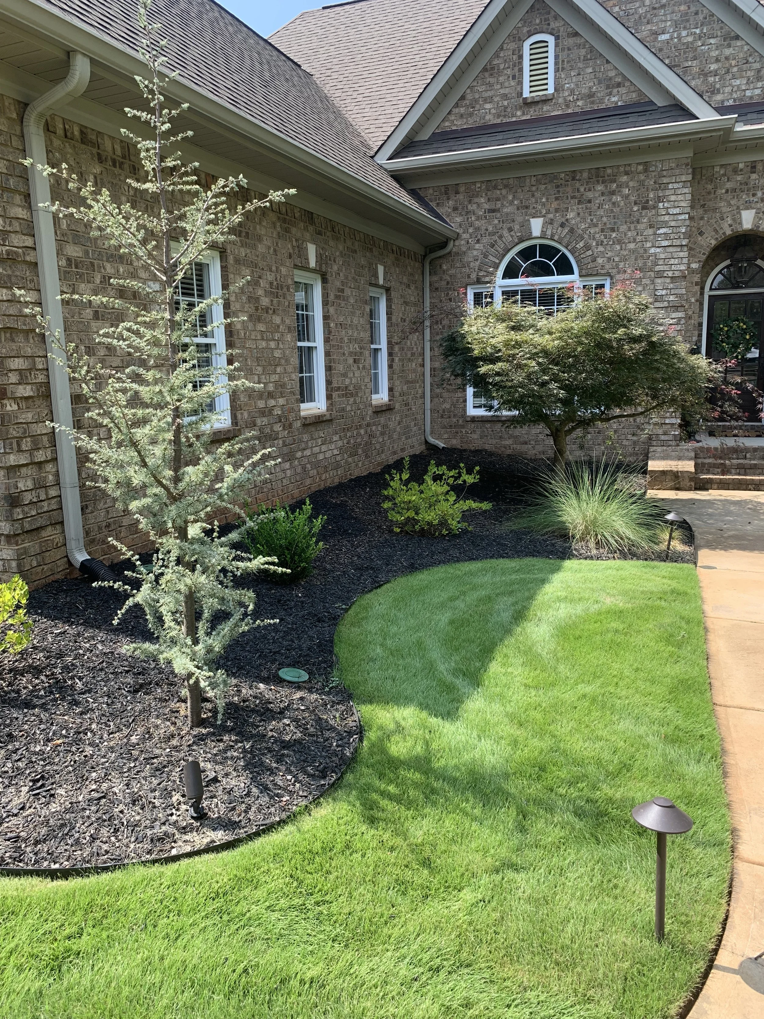 Well-maintained front yard with green lawn, small trees, shrubs, mulch beds, solar landscape lights, and a brick house with multiple windows and a staircase leading to the entrance.