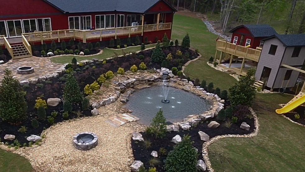 A landscaped backyard with a pond, rocks, trees, and a stone pathway, adjacent to red and gray houses with decks and a yellow slide.