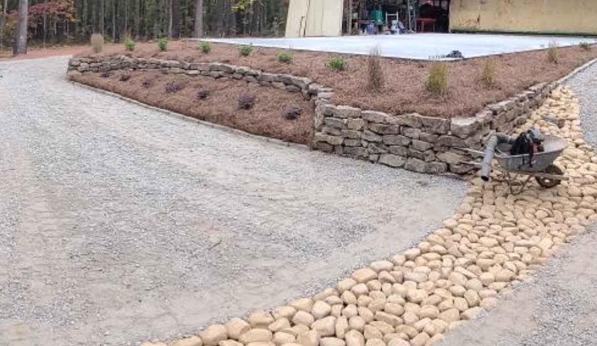 Landscape with gravel pathway, raised garden bed with rocks, small plants, and a wheelbarrow with gardening tools.