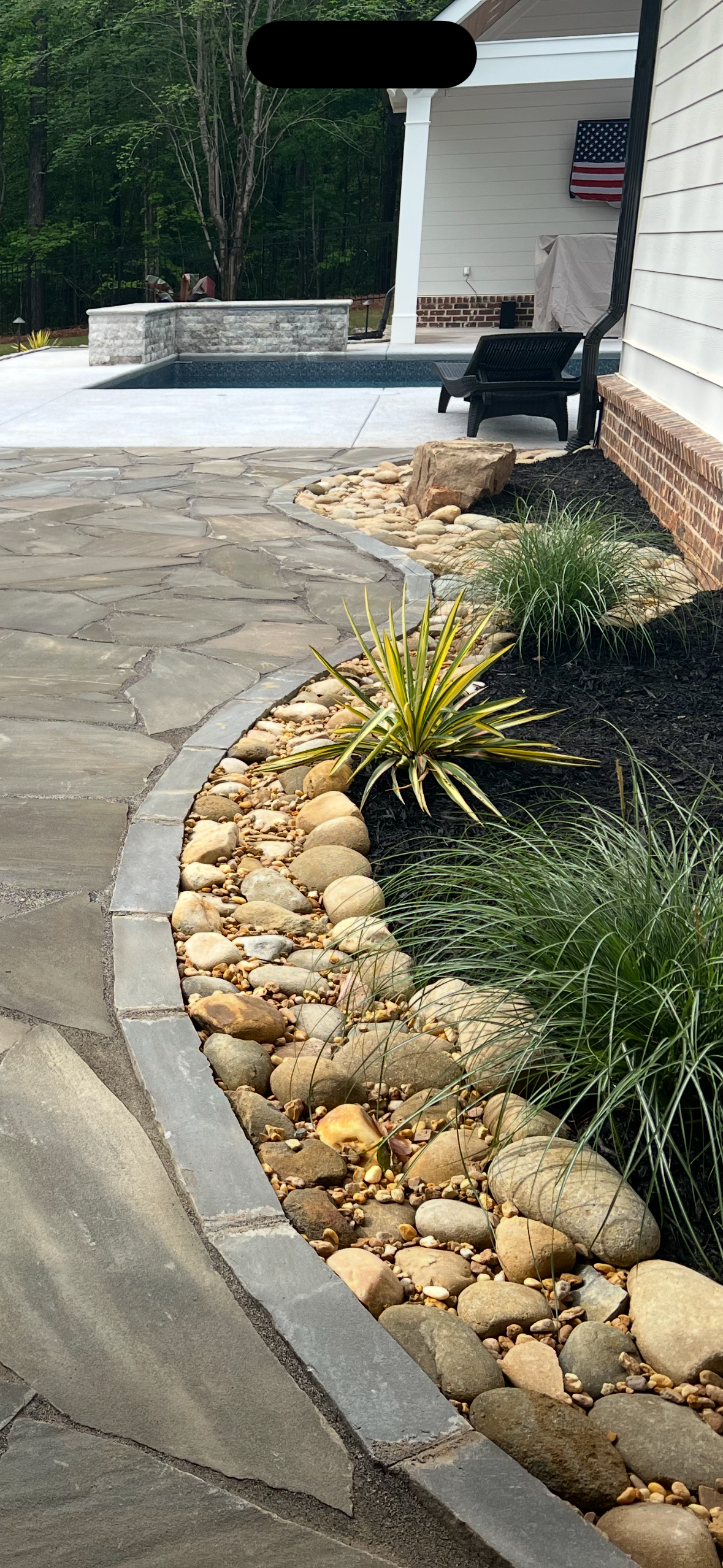 A backyard with a stone pathway, landscaping with rocks and plants, a small pool, and a house with an American flag.