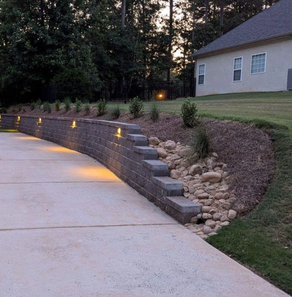A landscaped backyard terraced with a curved brick retaining wall illuminated by embedded lights, with small shrubs and rocks along the slope, neighboring a house with a lawn, trees, and a gate at sunset.