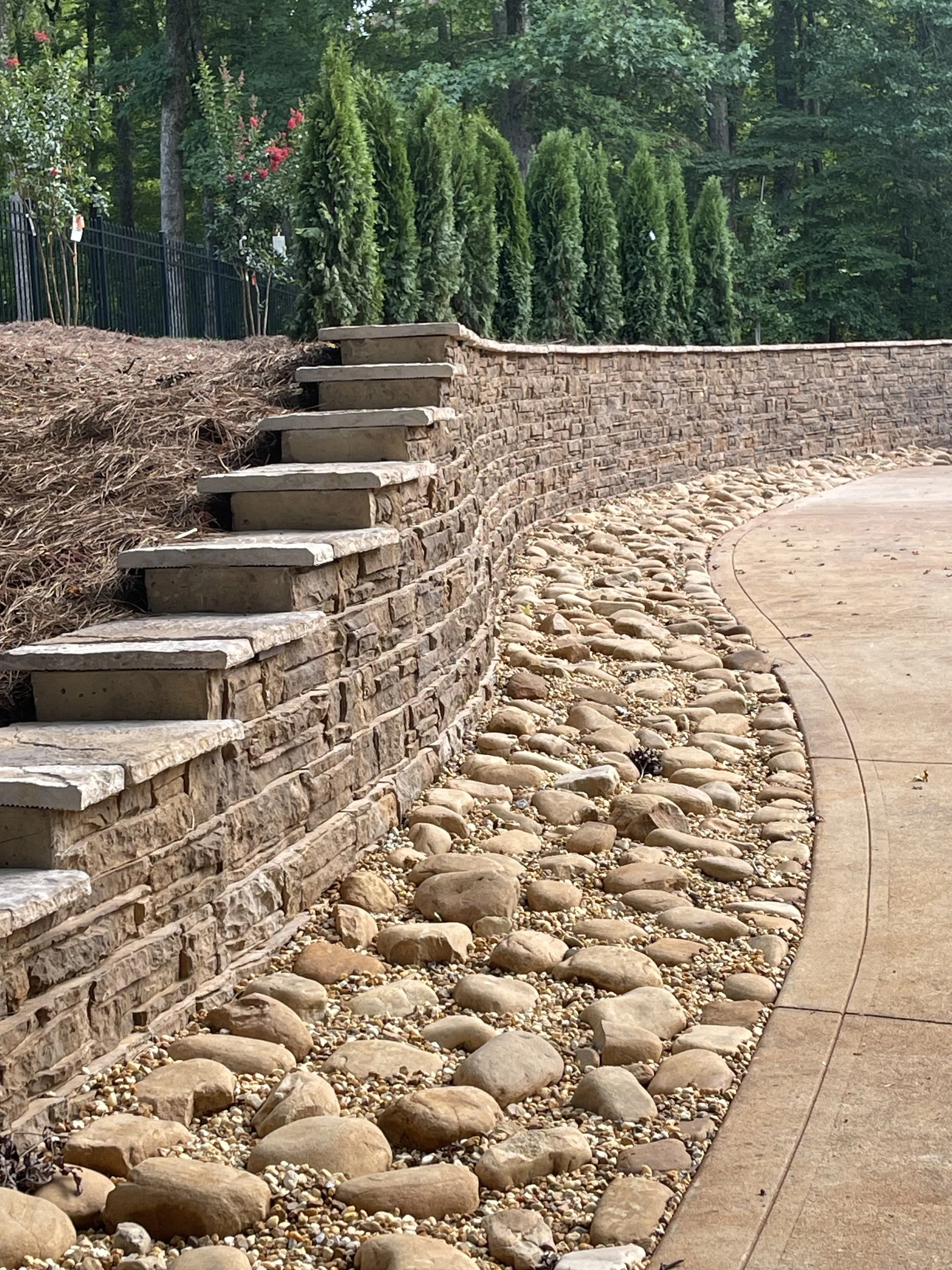 Curved stone pathway with boulder edging, stone steps, and a stone and wood retaining wall, surrounded by greenery.