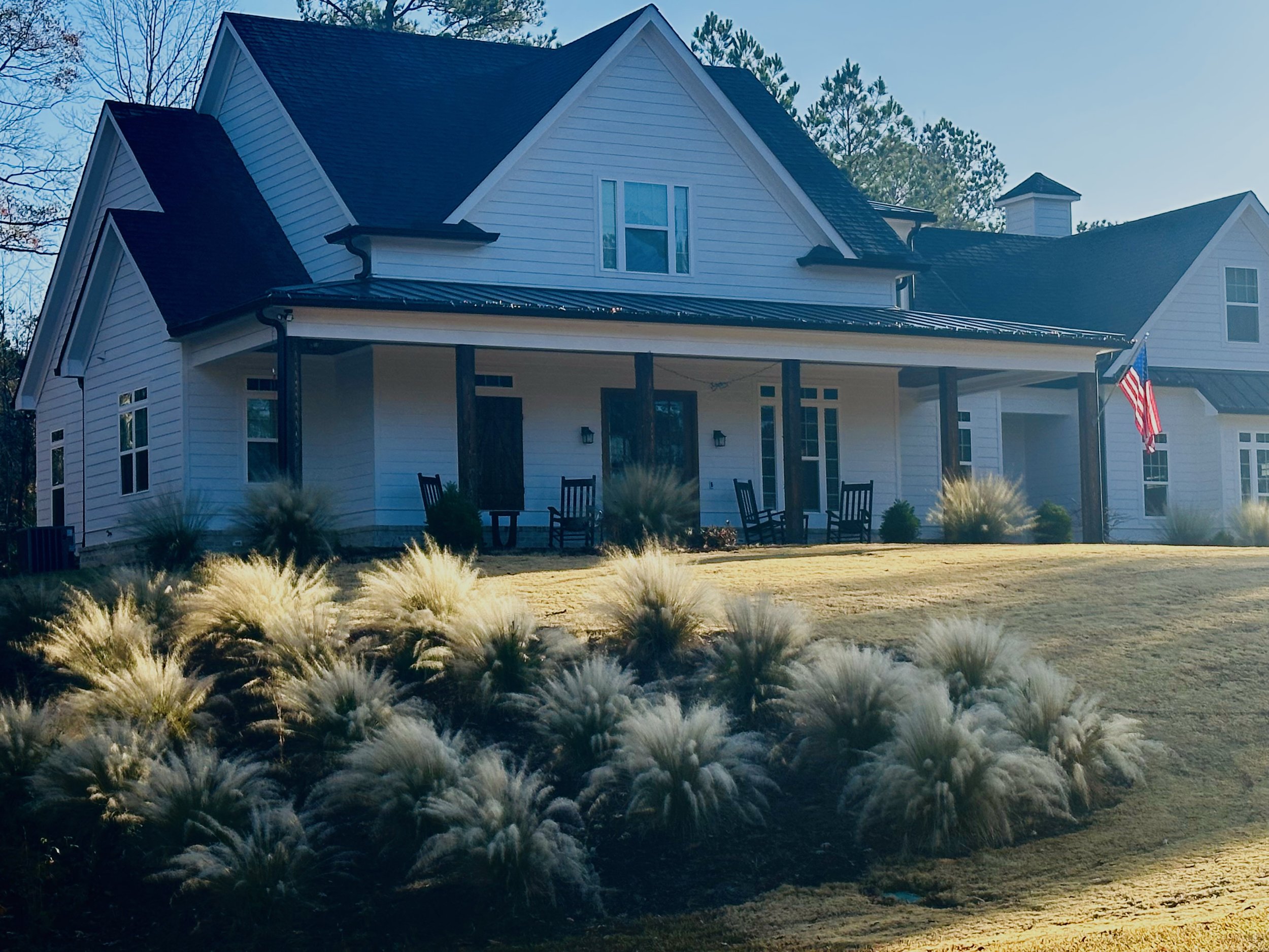 A white house with a dark roof and a large front porch with three rocking chairs; an American flag is flying on the right side; landscaped lawn with ornamental grasses in the foreground.