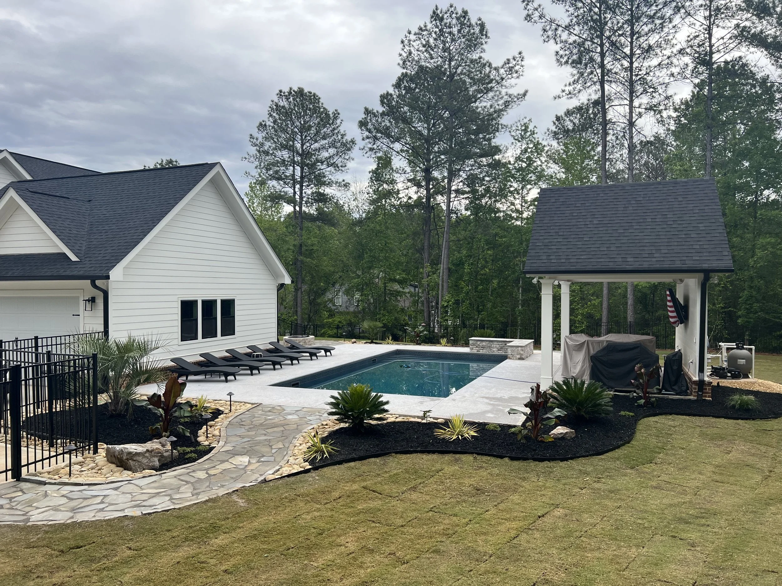Backyard with a rectangular swimming pool, black lounge chairs along the poolside, a small stone pathway, a white house with a black roof, a wooden shed with an American flag, surrounded by trees and greenery.
