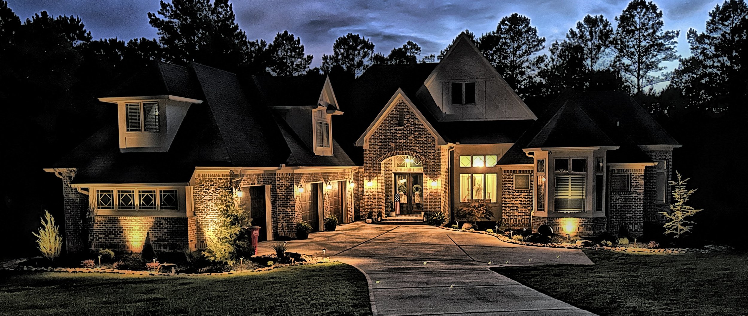 A large brick house illuminated by outdoor lights at dusk, with a concrete driveway, landscaped yard, and a wooded background.