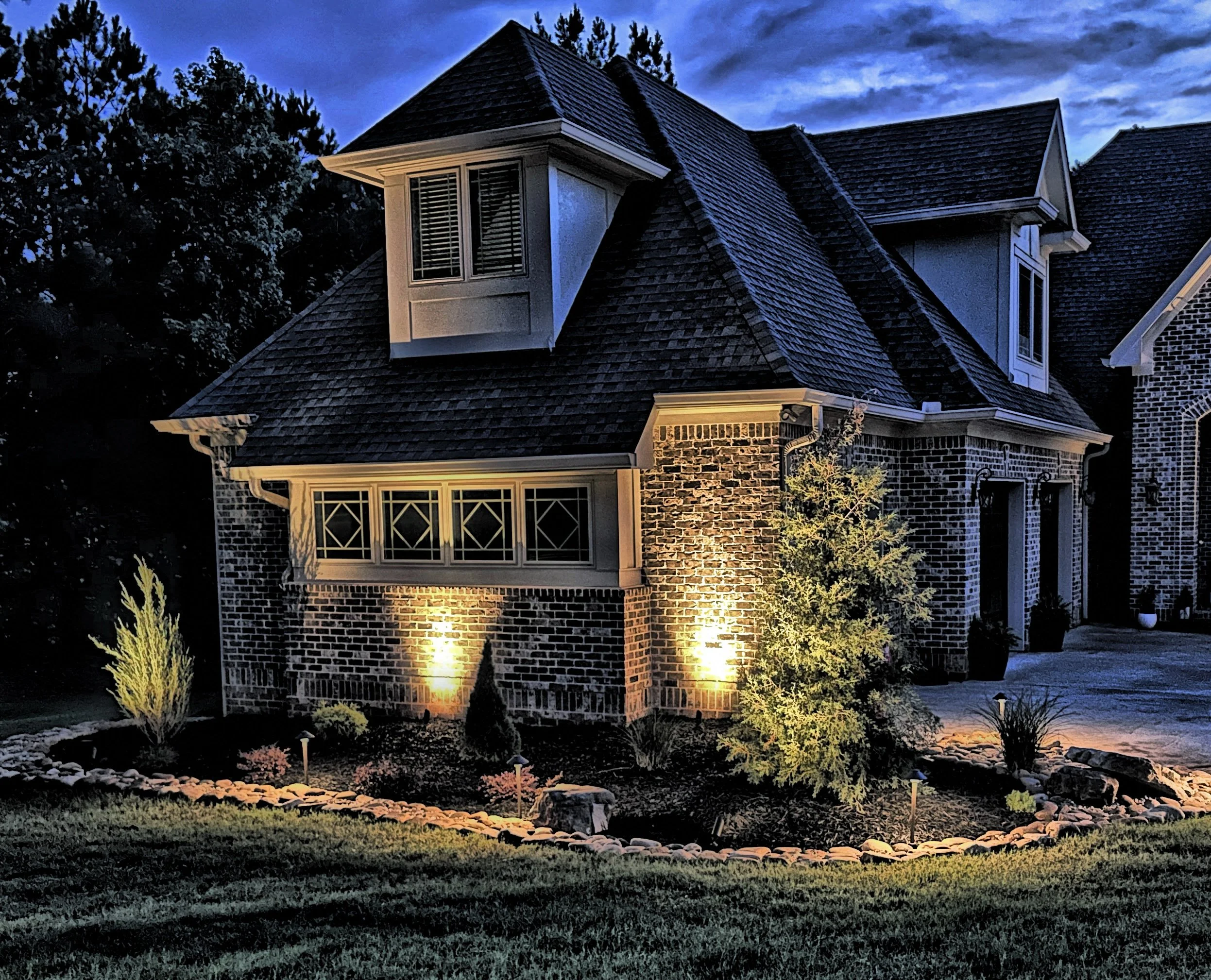 A house with brick walls illuminated by outdoor lights at dusk, with a landscaped front yard featuring bushes and small trees.