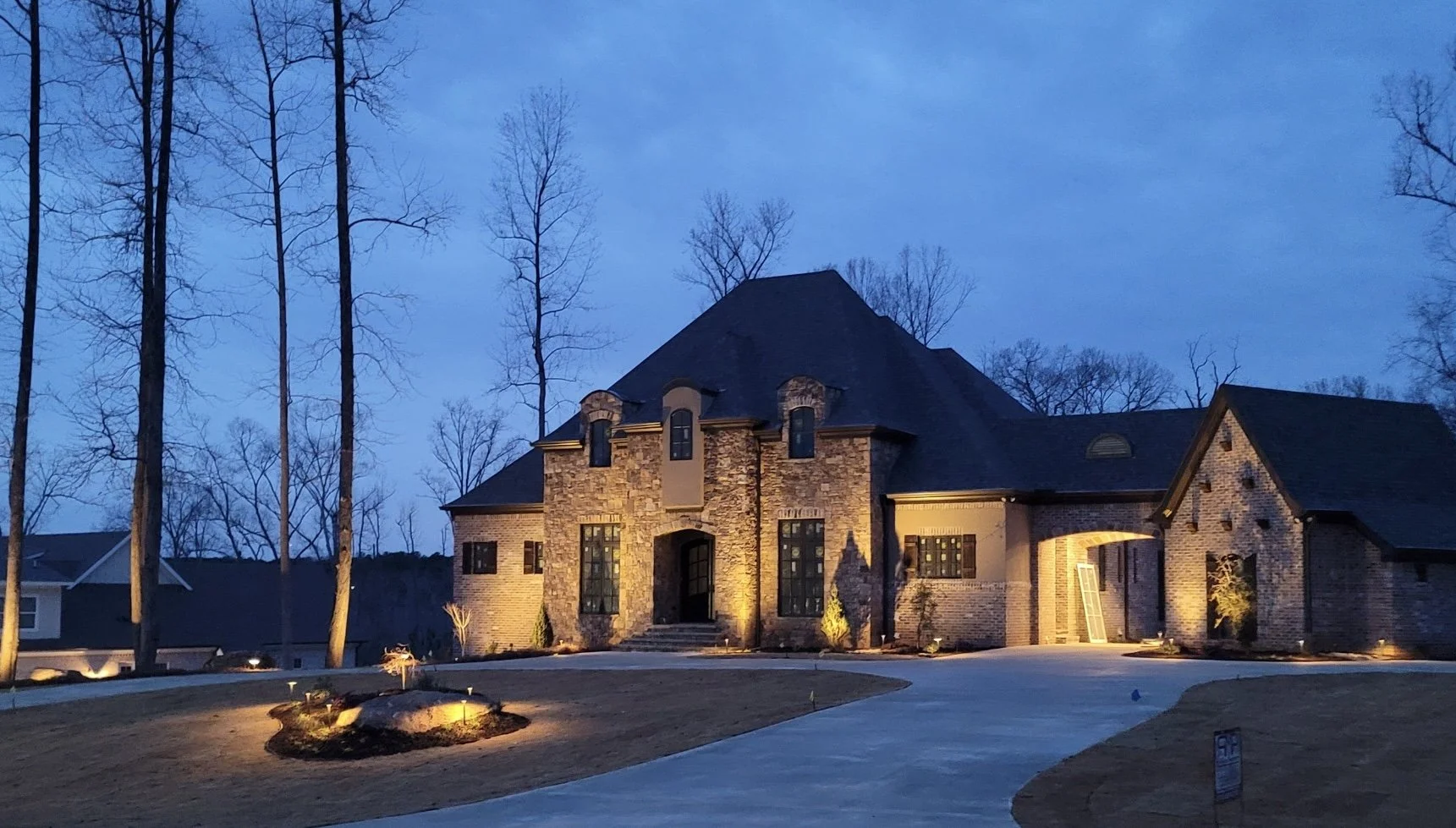Luxury brick house illuminated at dusk with a curved driveway and landscaped yard, leafless trees in the background.
