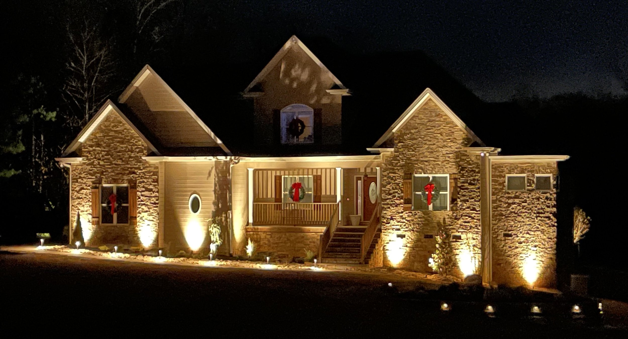 Nighttime view of a house decorated with Christmas wreaths and lights, with outdoor spotlights illuminating the exterior.