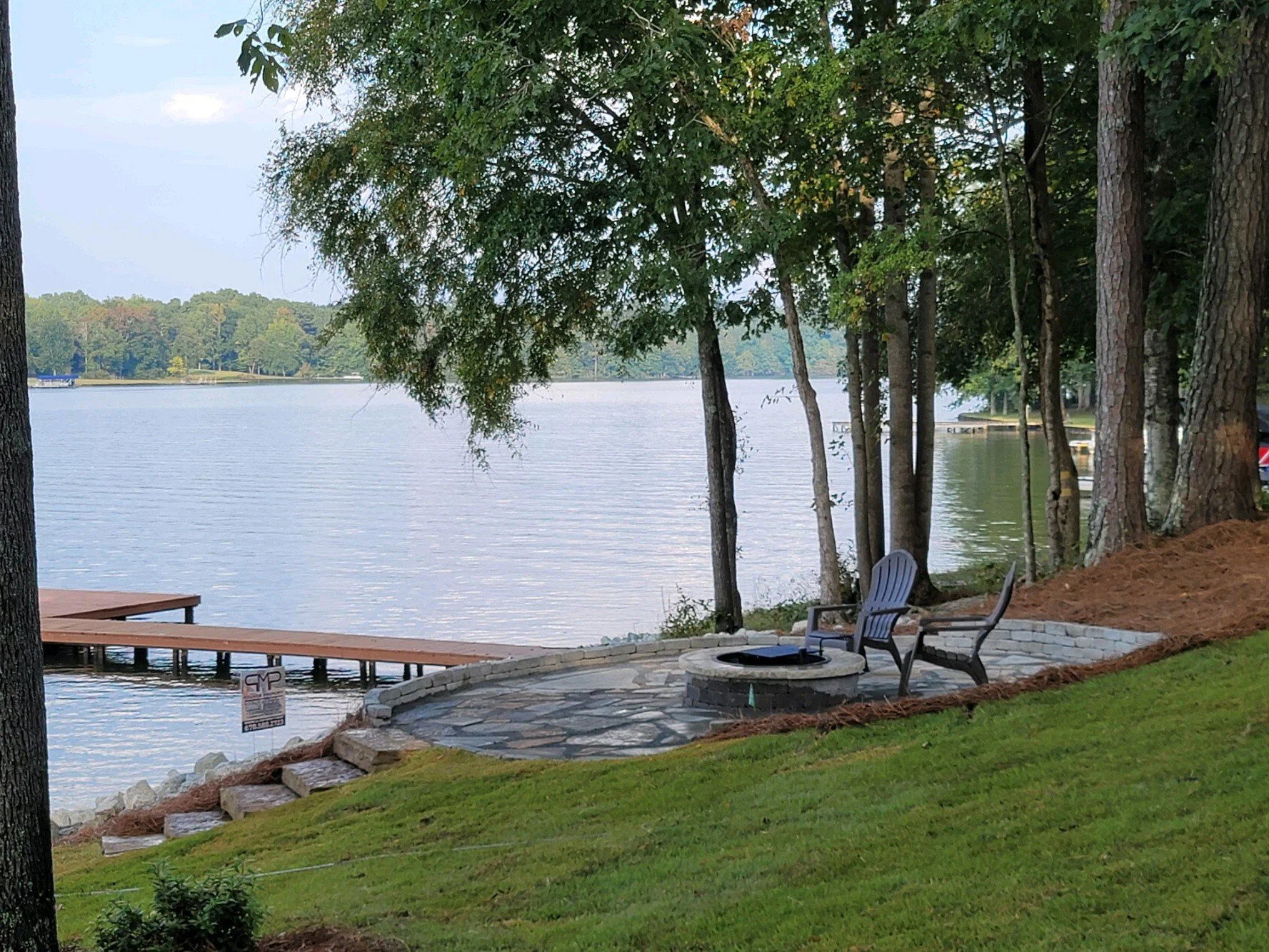 A lakeside scene featuring a stone patio with chairs around a fire pit, a grassy slope, and a wooden dock extending into the water, with trees lining the shore and a calm lake under a partly cloudy sky.
