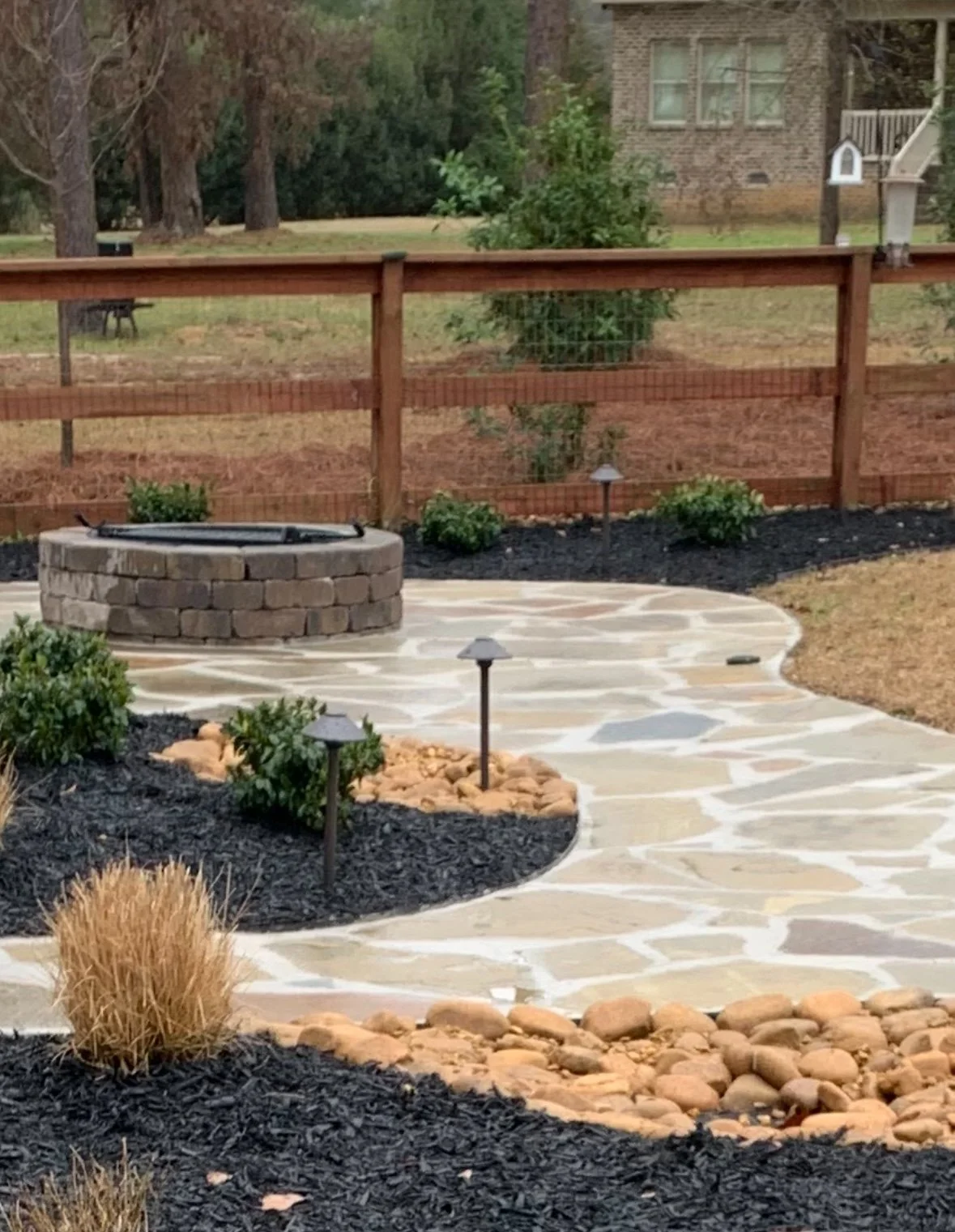 Backyard patio with flagstone pathway, a small brick fire pit, landscape lighting, and a wooden fence, with greenery and a house in the background.
