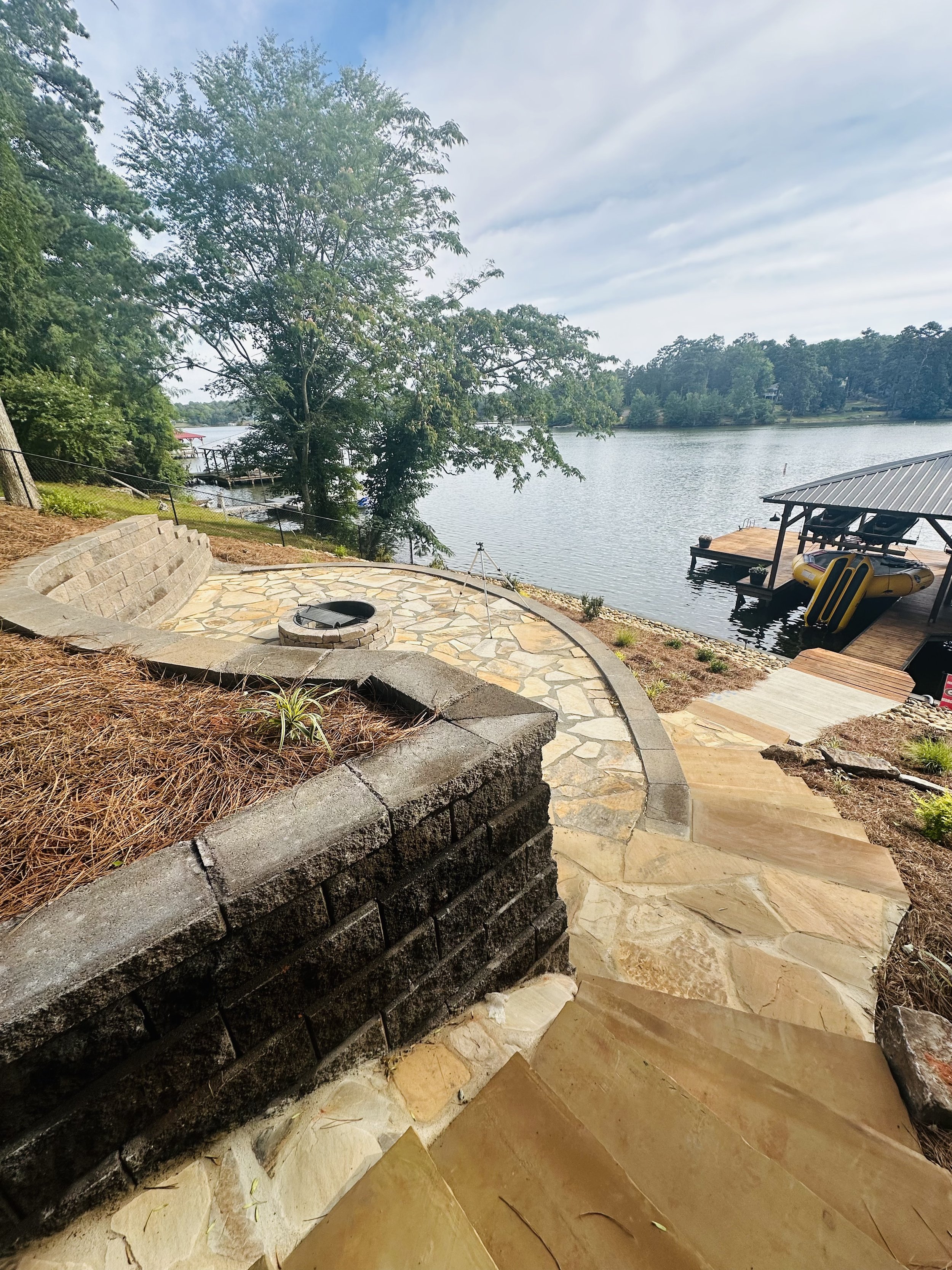 View of a lakeside patio with a stone pathway, fire pit, and a dock with a boat lift and inflatable boat, surrounded by trees and water.