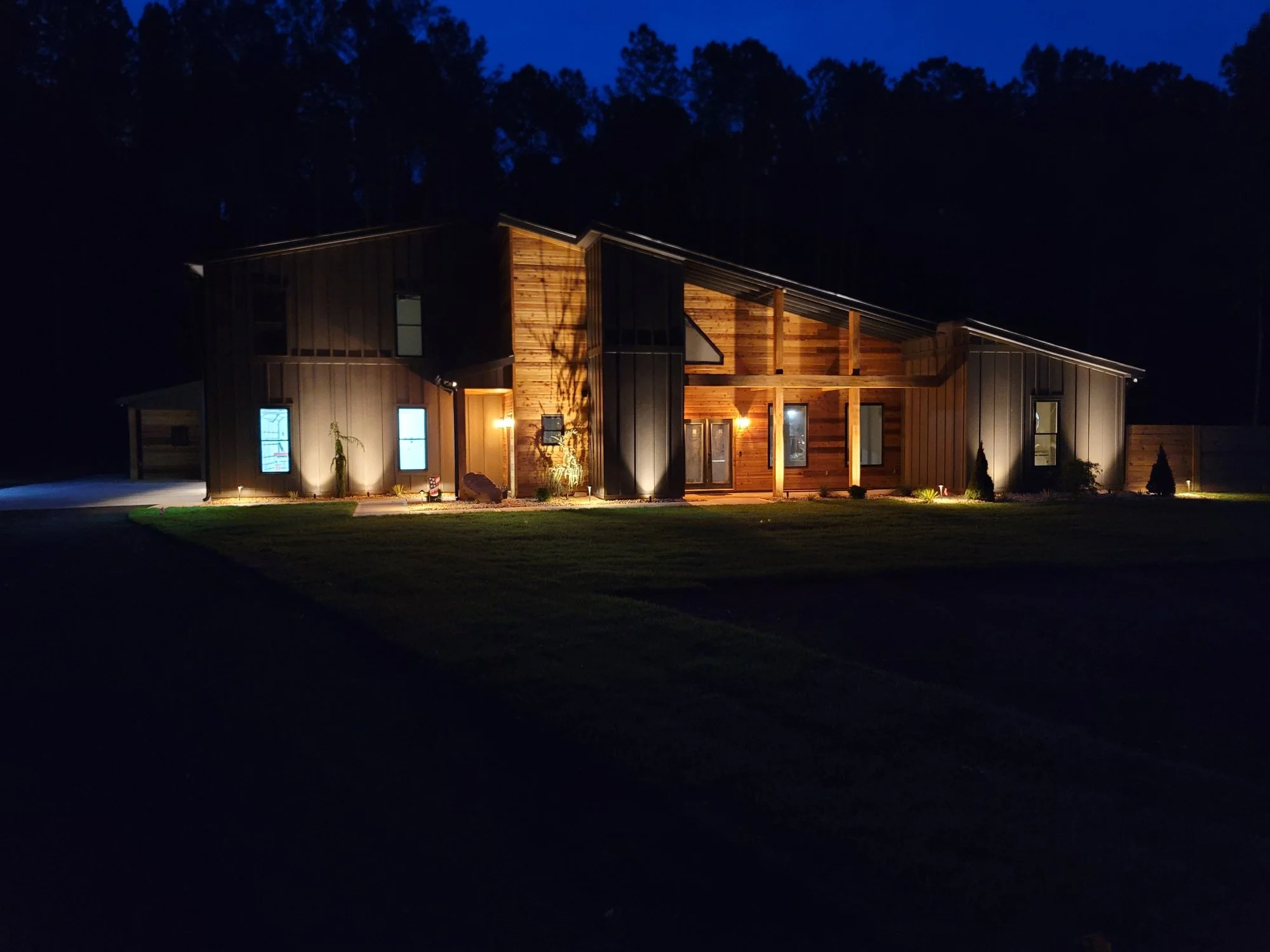 Nighttime photo of a modern house with wooden and metal exterior, illuminated windows and lights on the porch, with forested hill in the background.