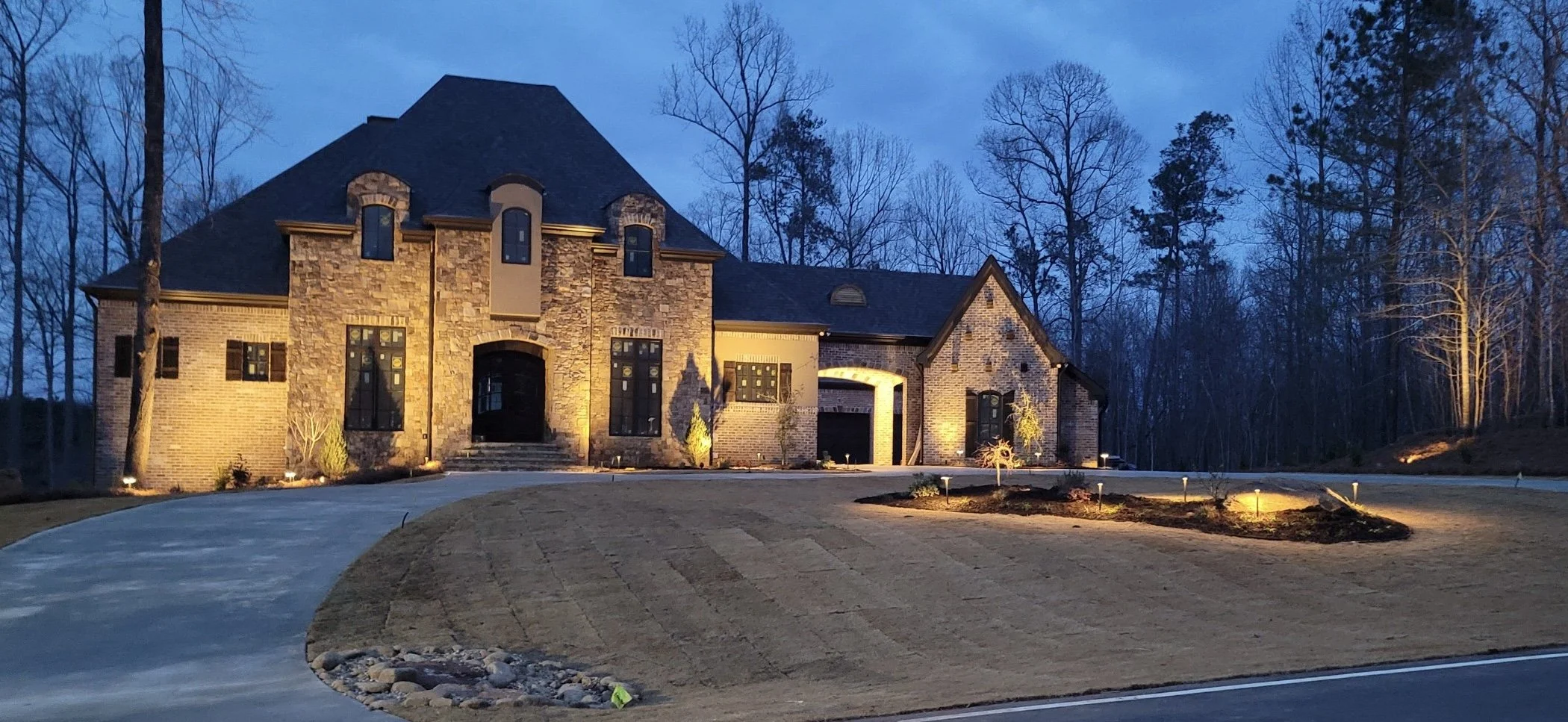 A large brick house with stone accents illuminated at dusk, featuring a curved driveway, landscaped yard with small trees, and tall trees in the background.