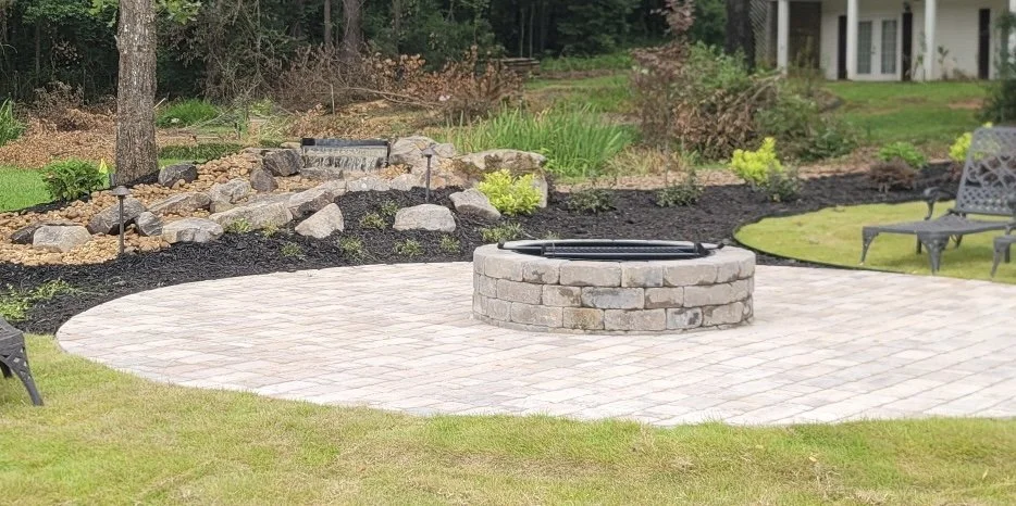 Backyard patio with a circular fire pit made of bricks, surrounded by a paved area, green lawn, garden beds, rocks, trees, and outdoor benches.