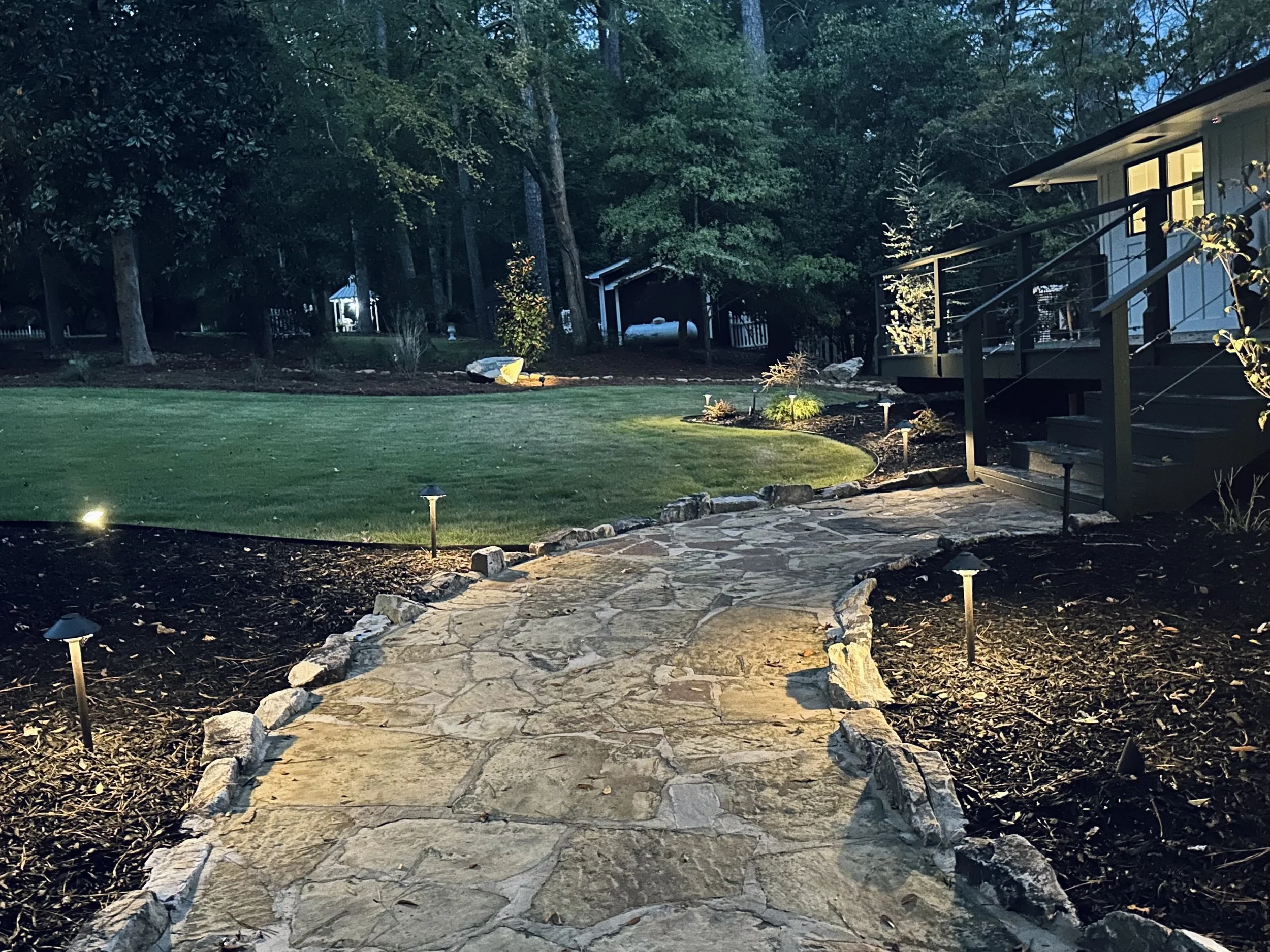 A stone pathway illuminated by garden lights leading to a house porch in a backyard with a well-maintained lawn and surrounding trees.