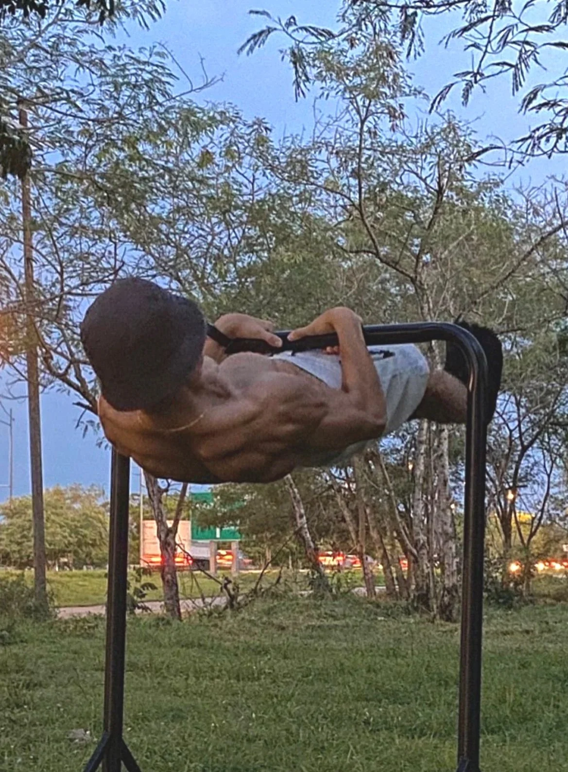 Un hombre sin camisa haciendo touch front lever, con árboles de fondo y un cielo parcialmente nublado.