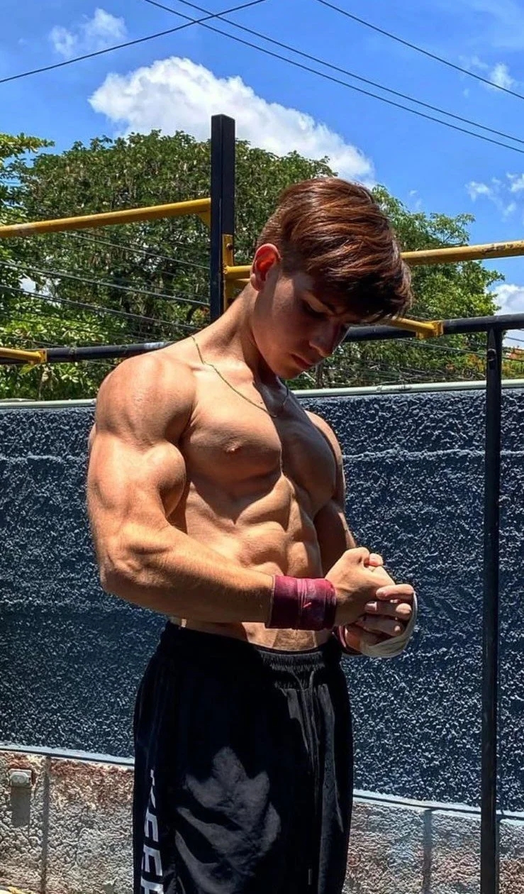 Joven en postura de entrenamiento al aire libre, sin camiseta, con pantalones deportivos negros, en un entorno con cielo azul y árboles.