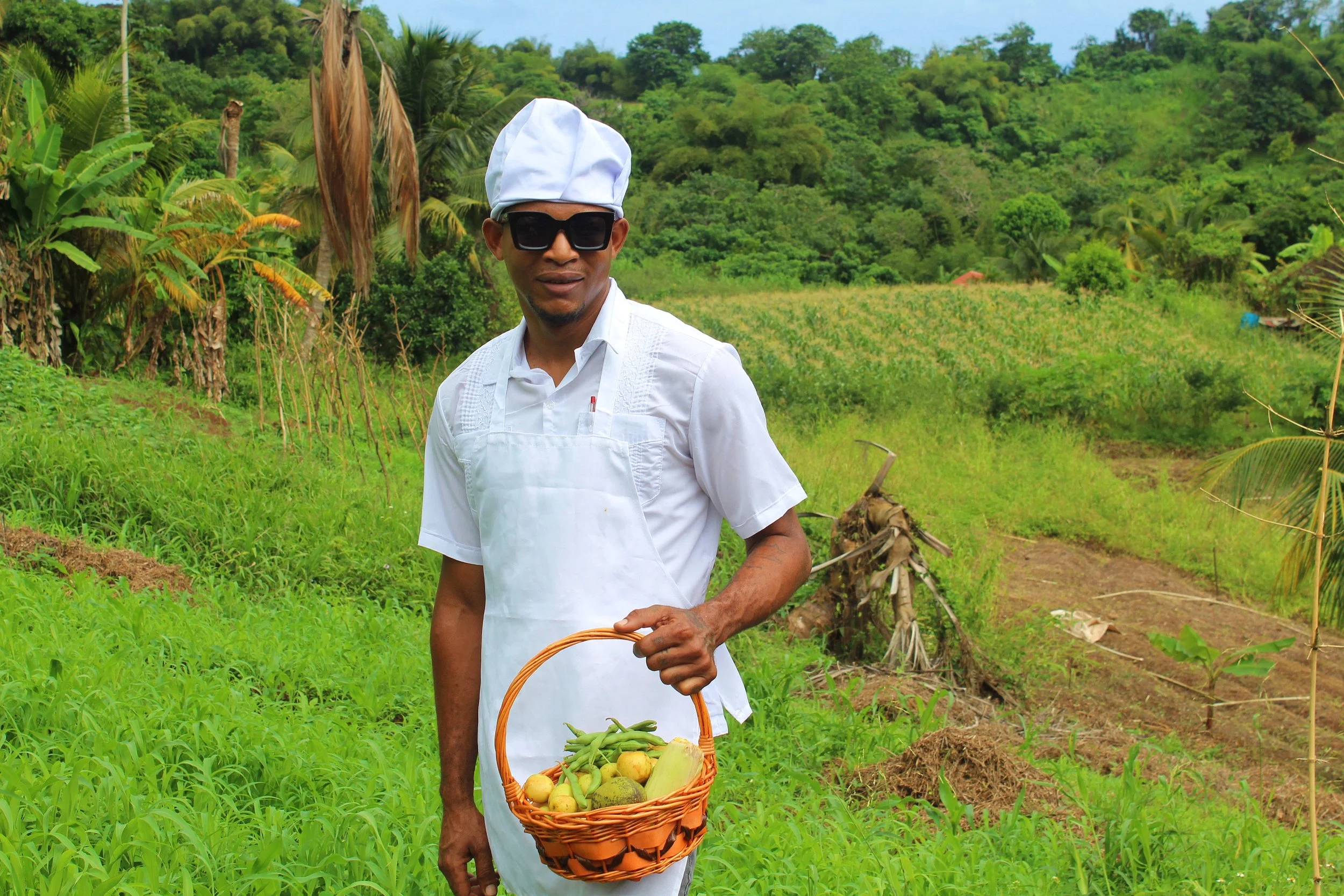 A man dressed as a chef in white with a chef's hat, sunglasses, and apron, standing outdoors in a lush green farm or garden, holding a basket of fresh vegetables.