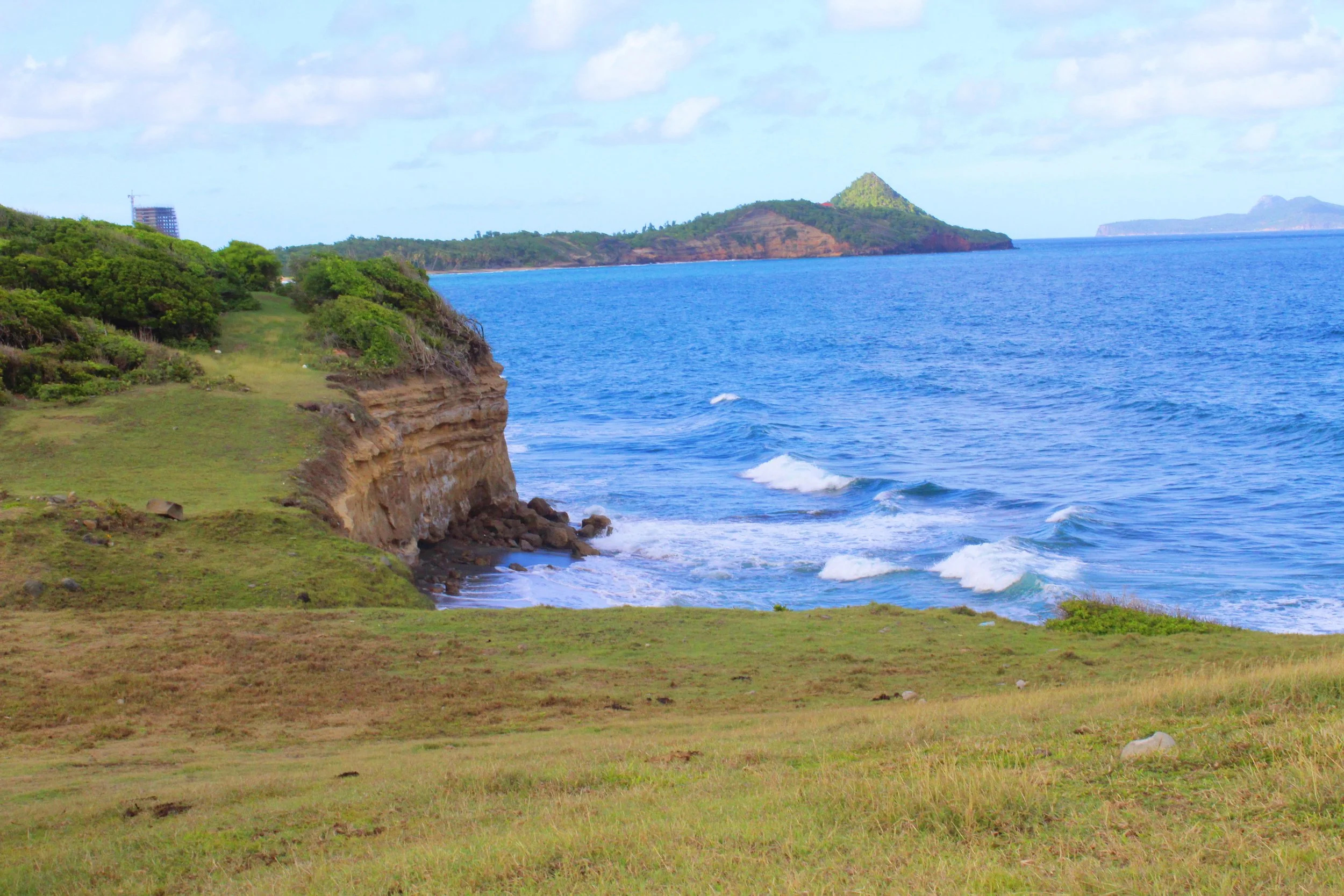 Seaside landscape with grassy cliffs, ocean waves, and distant islands under a partly cloudy sky.