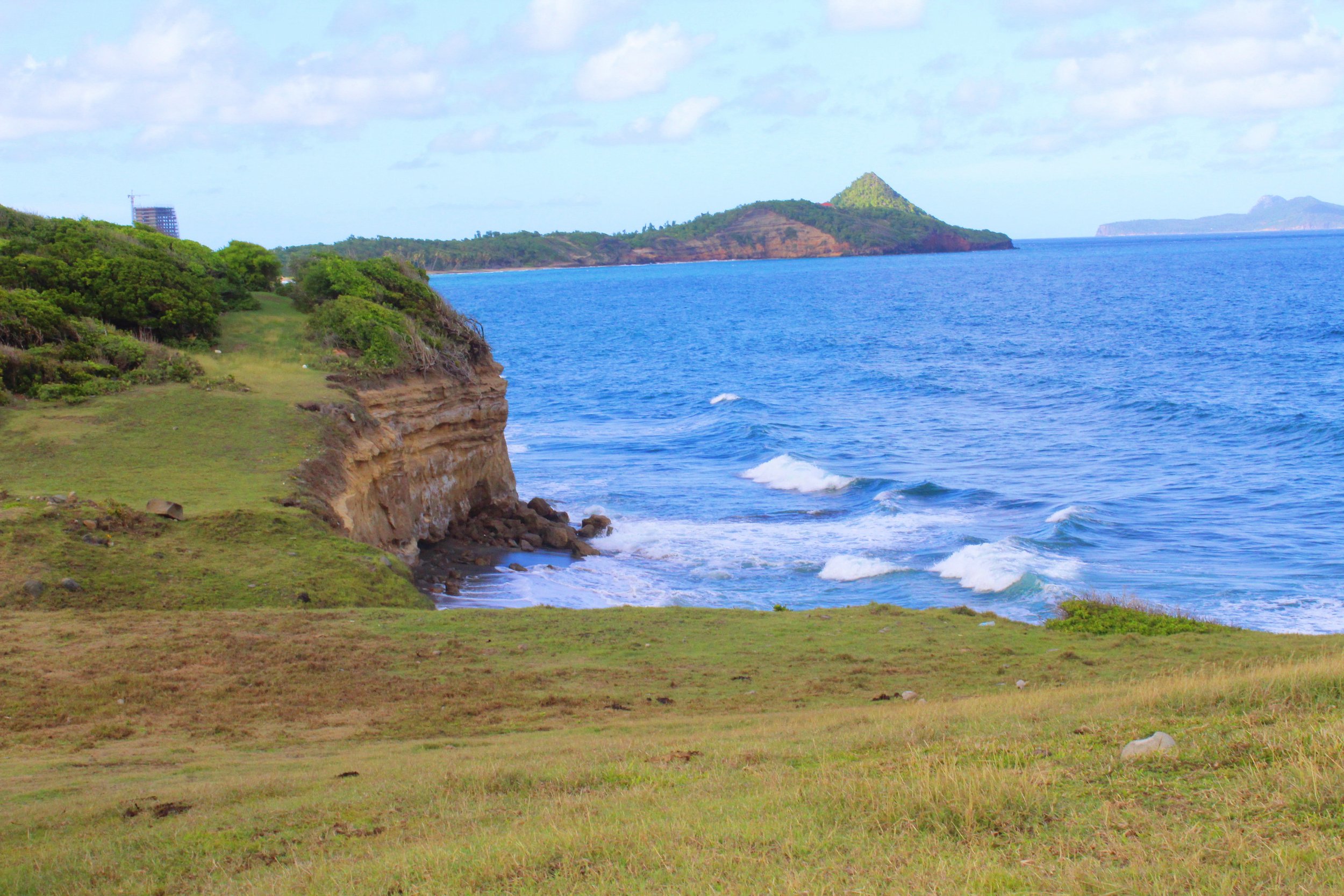 Scenic view of a coastal landscape with green grassy cliffs, blue ocean waves, and islands in the distance under a partly cloudy sky.