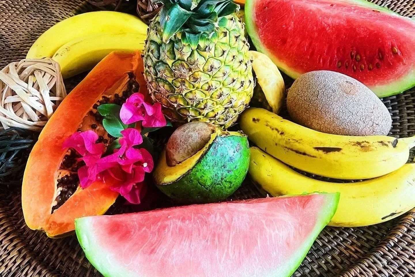 A variety of fresh tropical fruits including pineapple, bananas, watermelon, papaya, avocado, and a small flower, arranged on a woven surface.