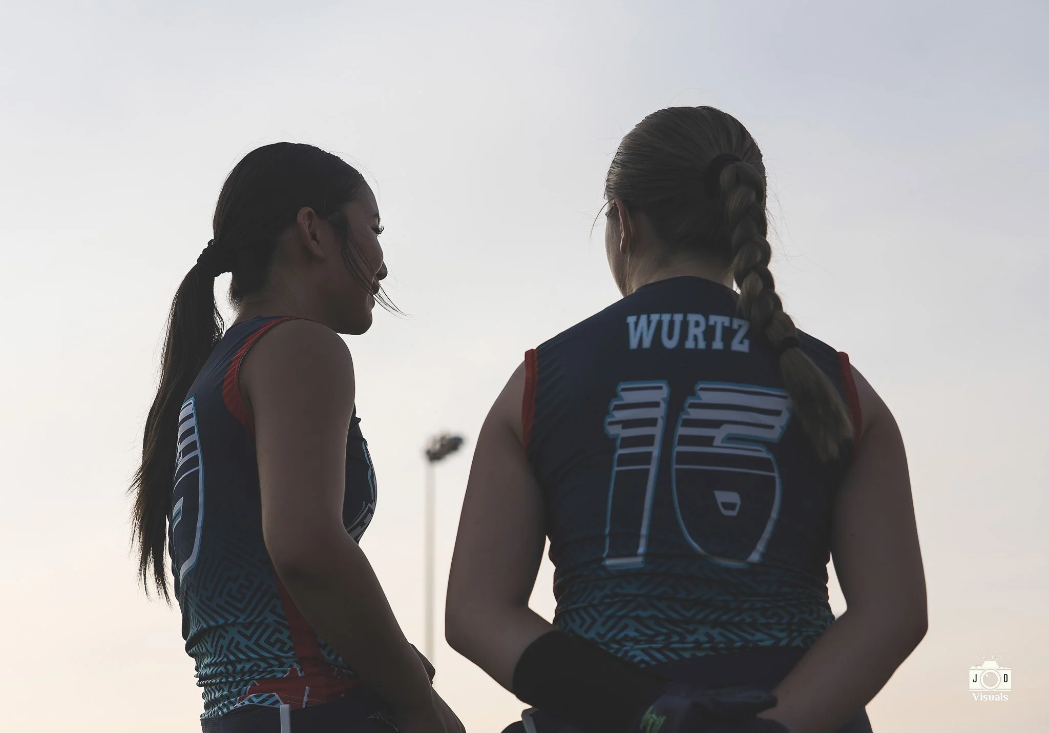 Two female athletes in sports uniforms talking outdoors during sunset, with a sports field light in the background.