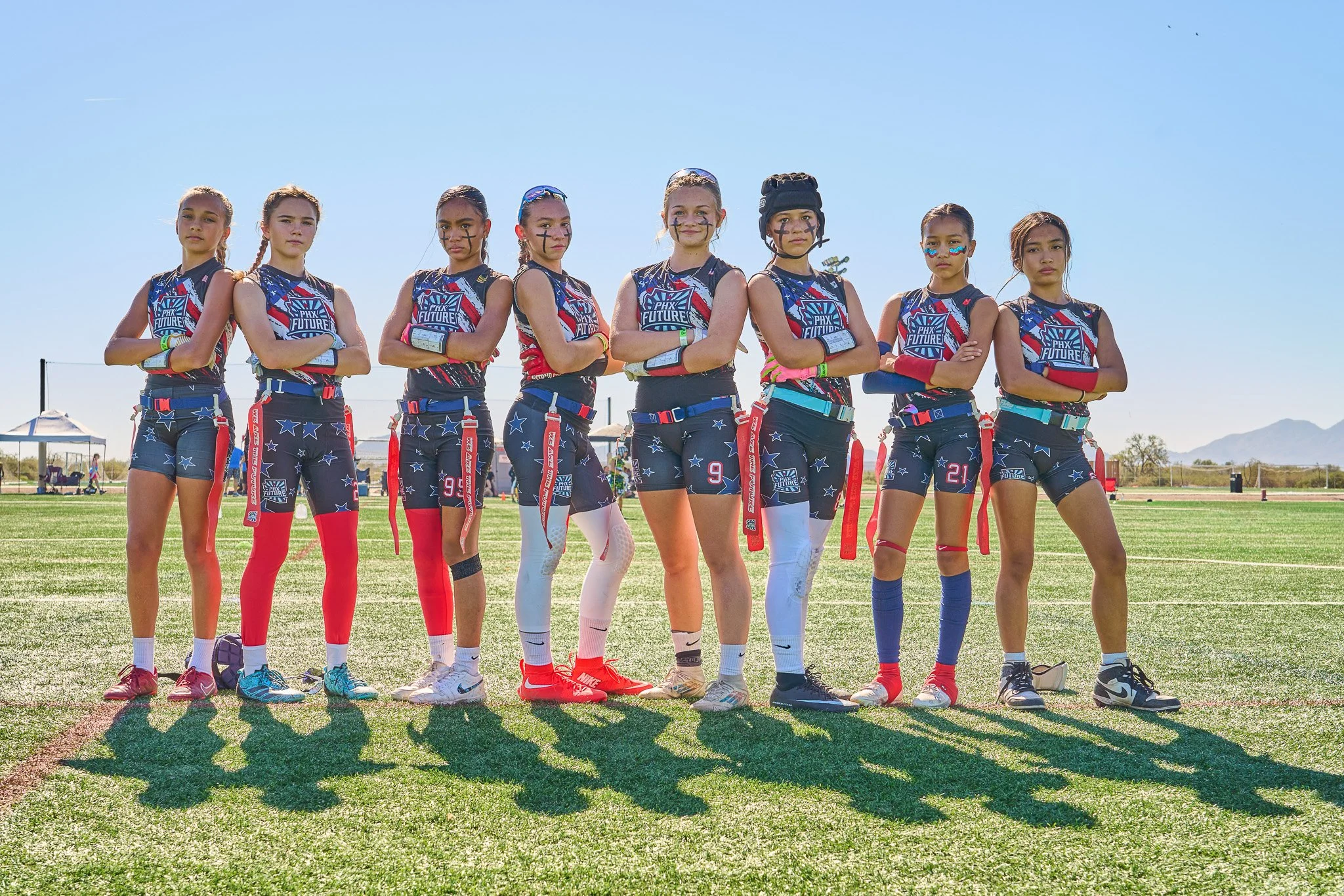 Eight young female flag football players standing shoulder to shoulder on a field, wearing matching team uniforms with arm and leg pads and face paint, under clear blue sky.