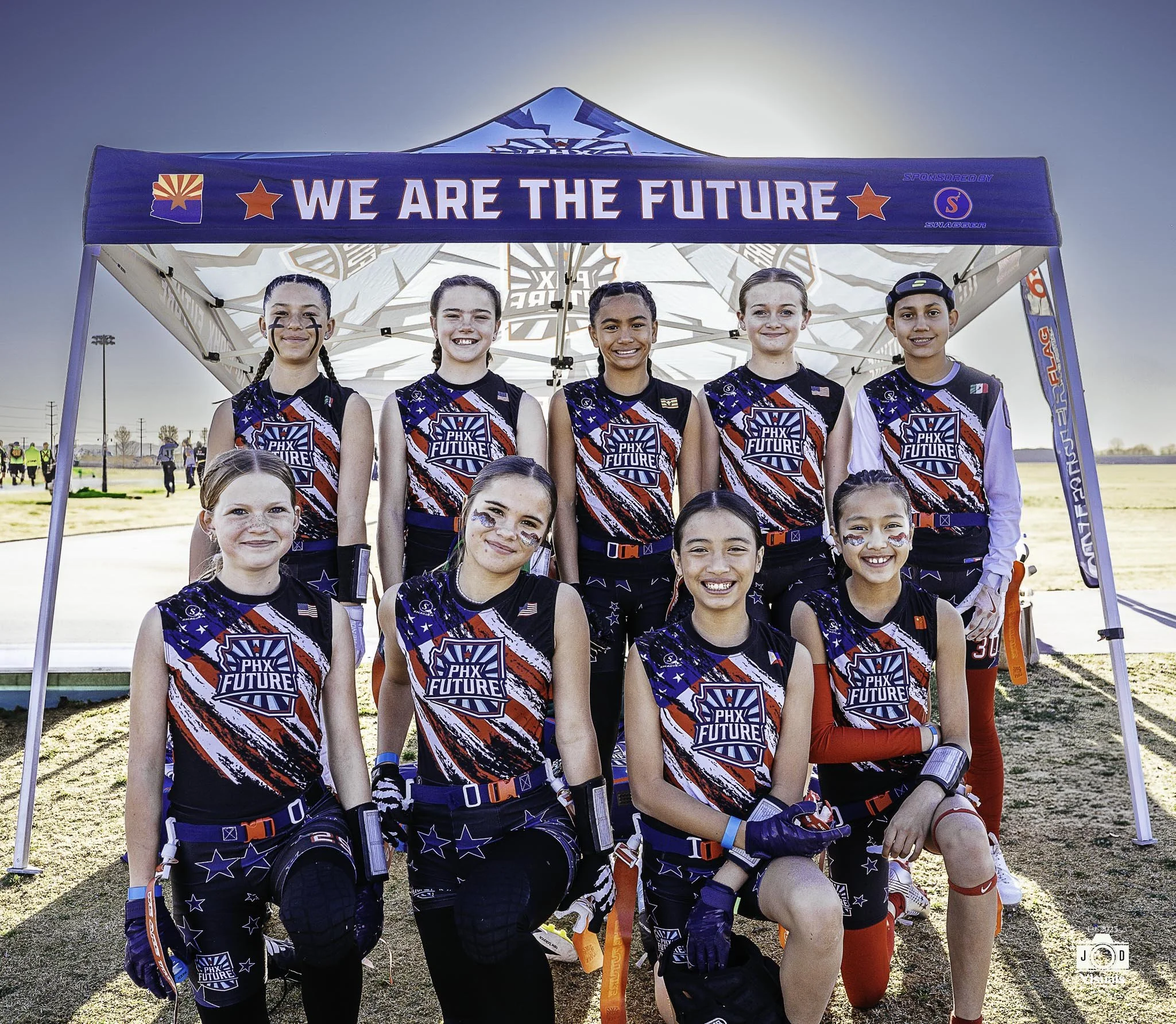 A girls' flag football team standing under a tent with the phrase 'We Are The Future' written on it, wearing matching jerseys with a patriotic design, smiling after a game or practice outdoors.