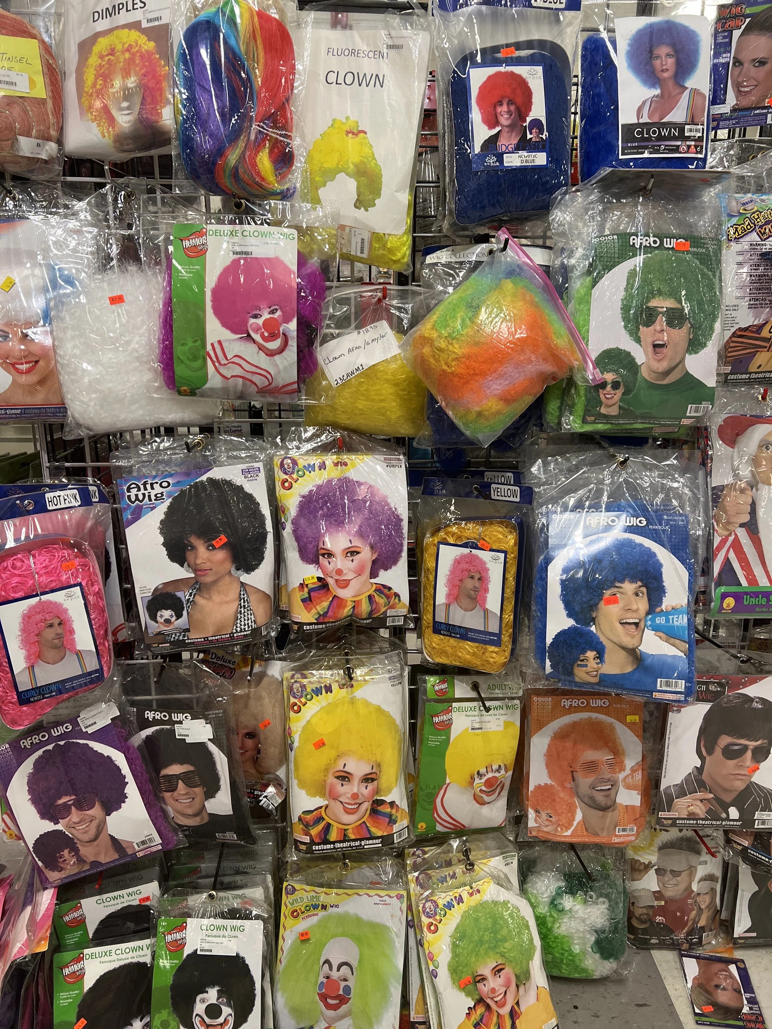 Display of colorful clown wigs and costume accessories on store hooks, featuring various styles and colors including pink, yellow, green, purple, blue, and rainbow, with clown face packaging and promotional pictures.