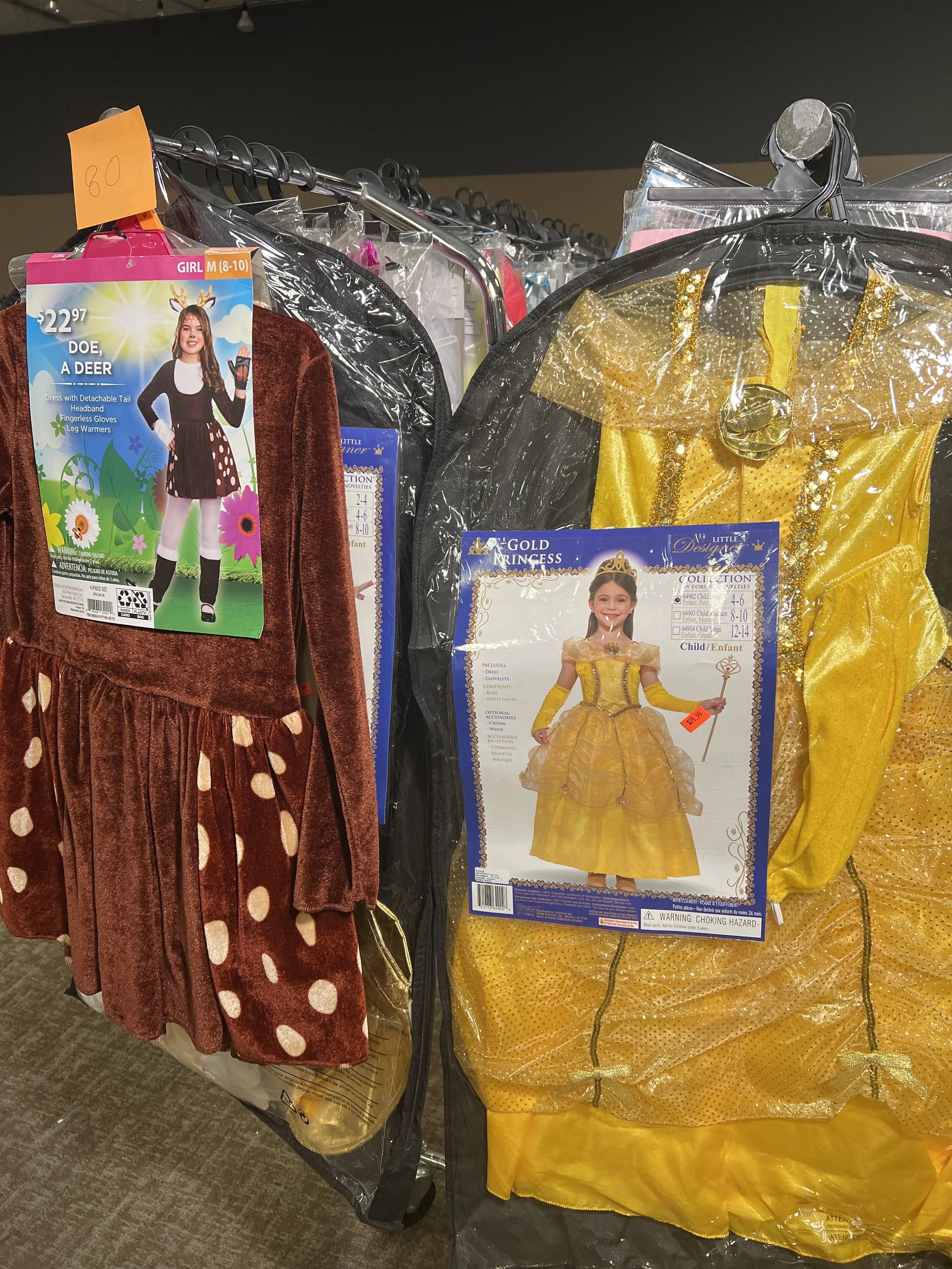Two children's costumes hanging on a rack, one is a girl dress with a detachable tail and reindeer headband, the other is a princess dress in yellow.