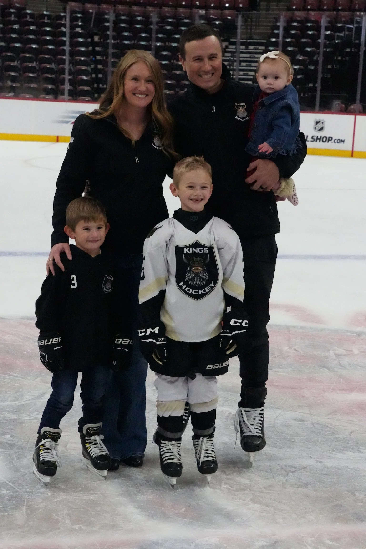 A family of five on an ice rink, two adults, two young children in hockey gear, and a toddler. They are smiling and posing for the photo.