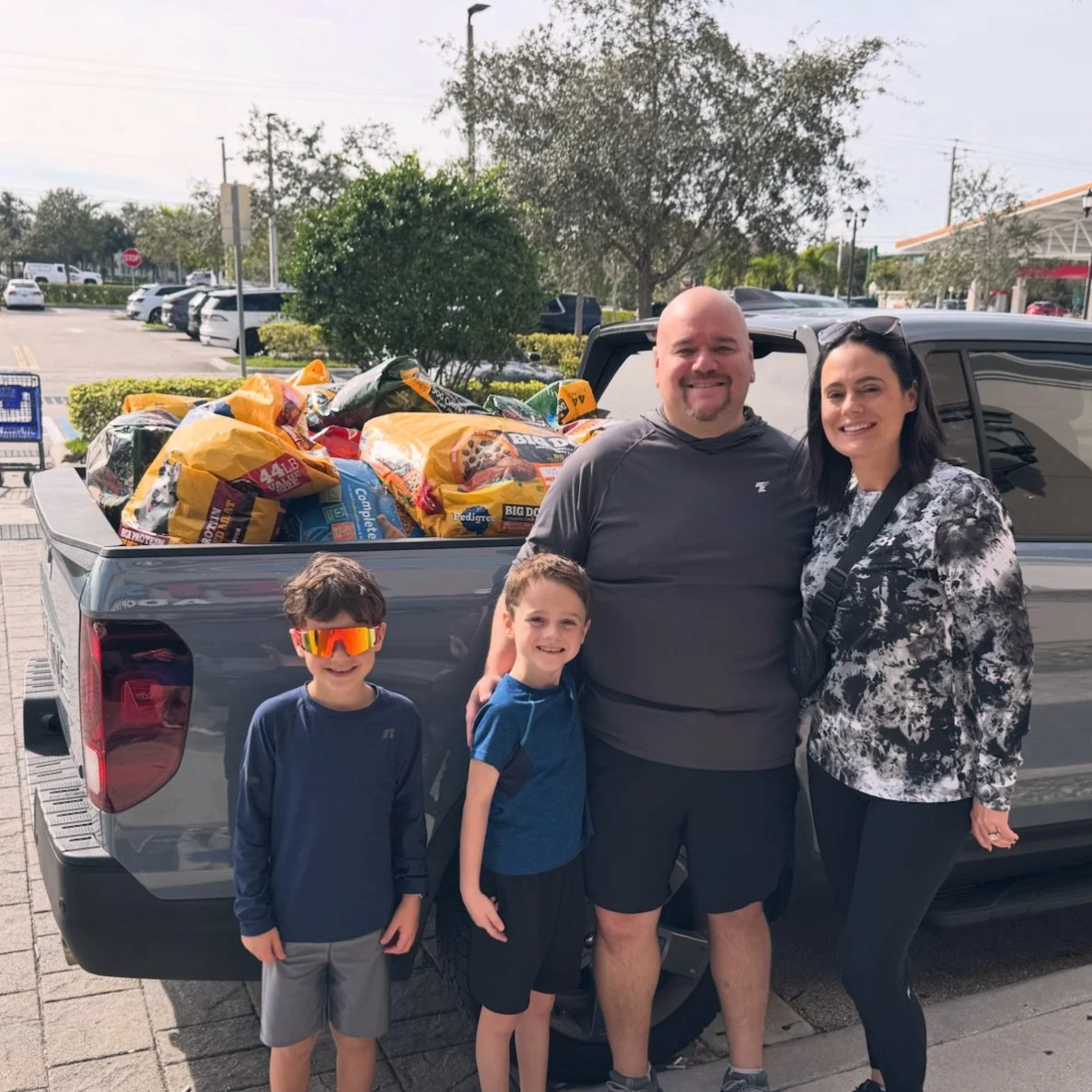 A family of four standing next to a pickup truck loaded with grocery bags in a parking lot.