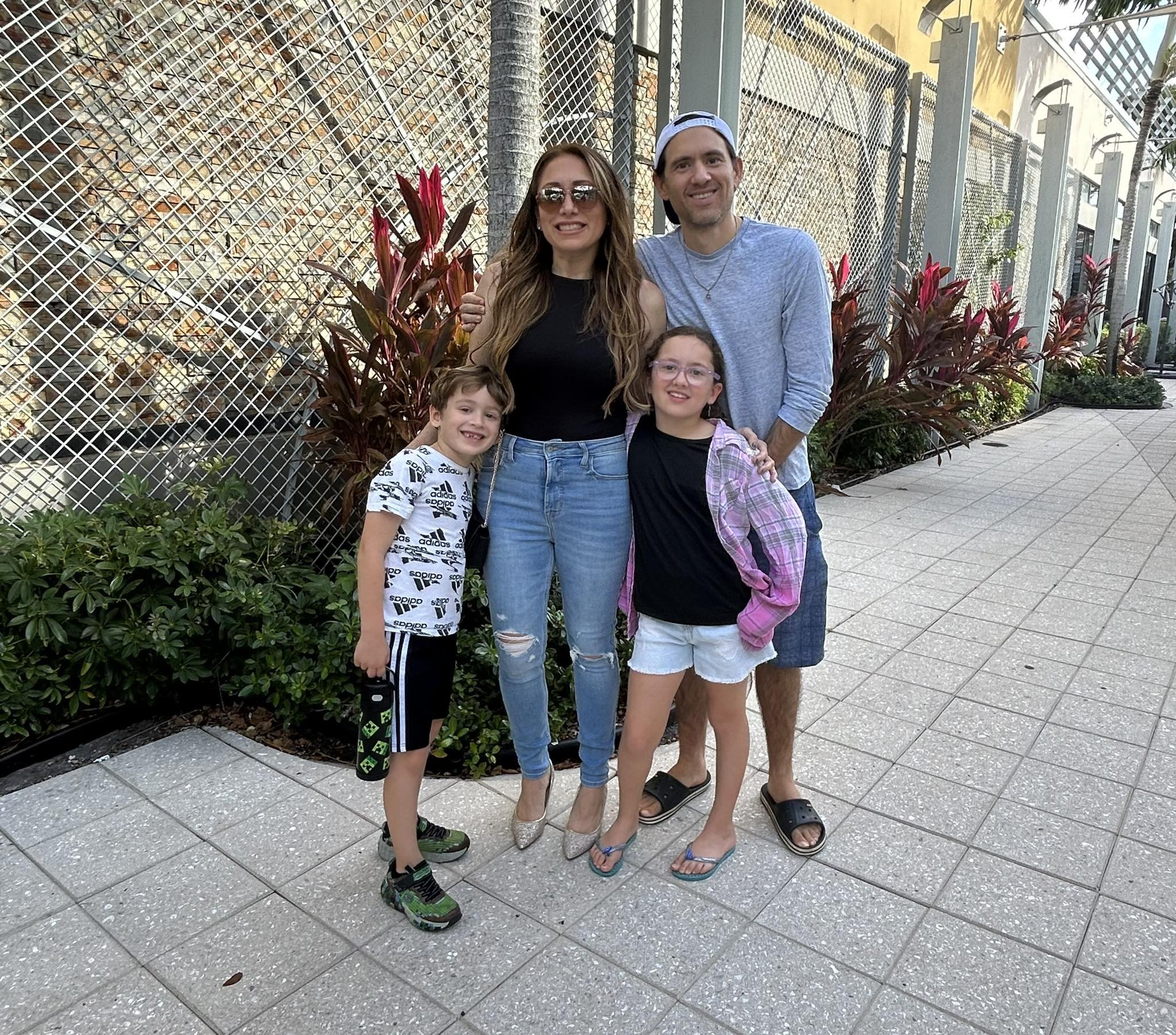 A family of four standing outdoors, with a woman and a man in the back and two children in front. The woman has long brown hair, sunglasses, a black top, and ripped jeans. The man has short dark hair, a backward white cap, a gray long-sleeve shirt, and shorts. The boy has short light brown hair, a white Adidas shirt, and athletic shorts. The girl has glasses, long brown hair, a black shirt, a pink plaid jacket, and white shorts.