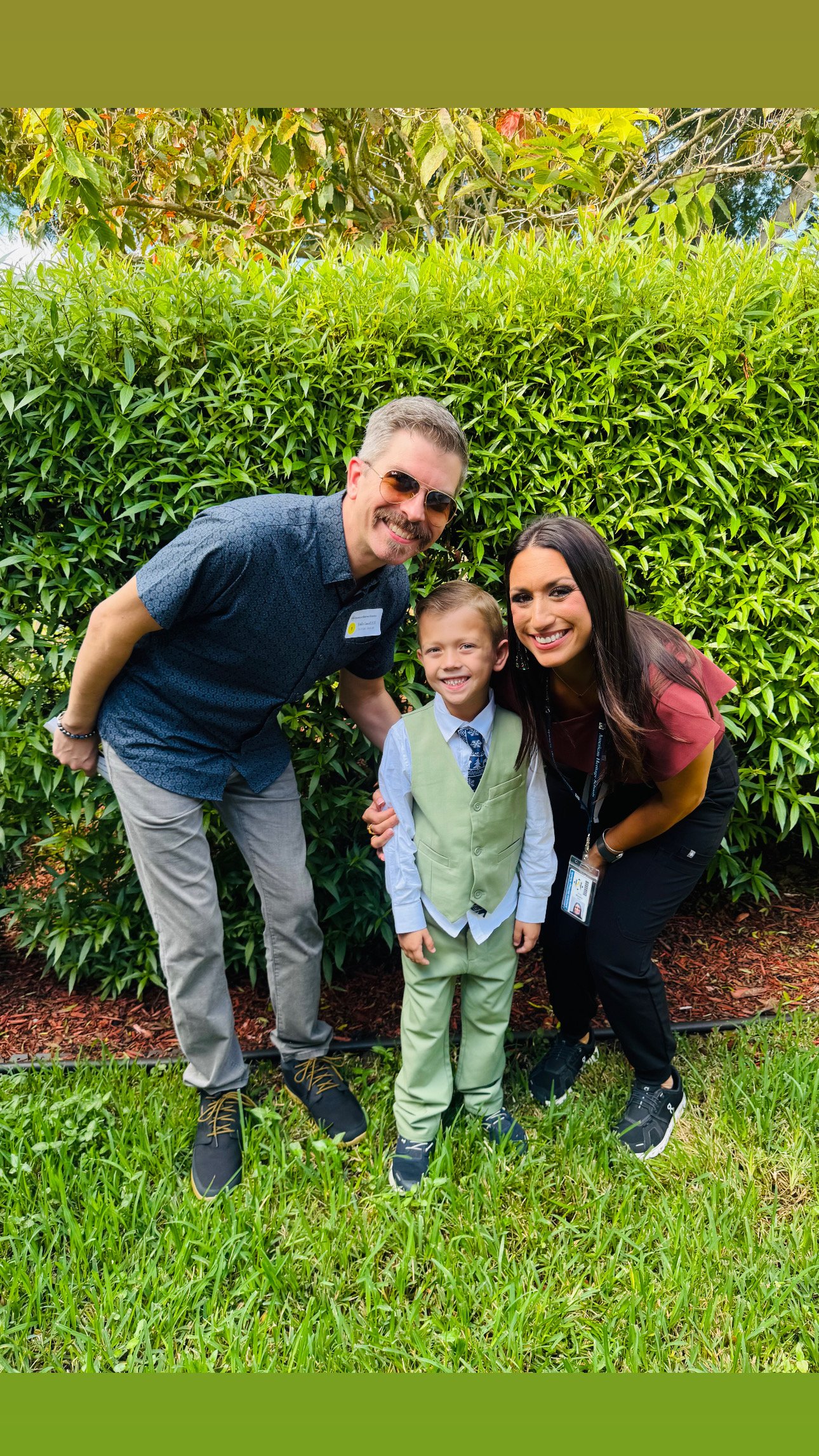 A man, woman, and young boy smiling and posing together outdoors in front of a green bush.