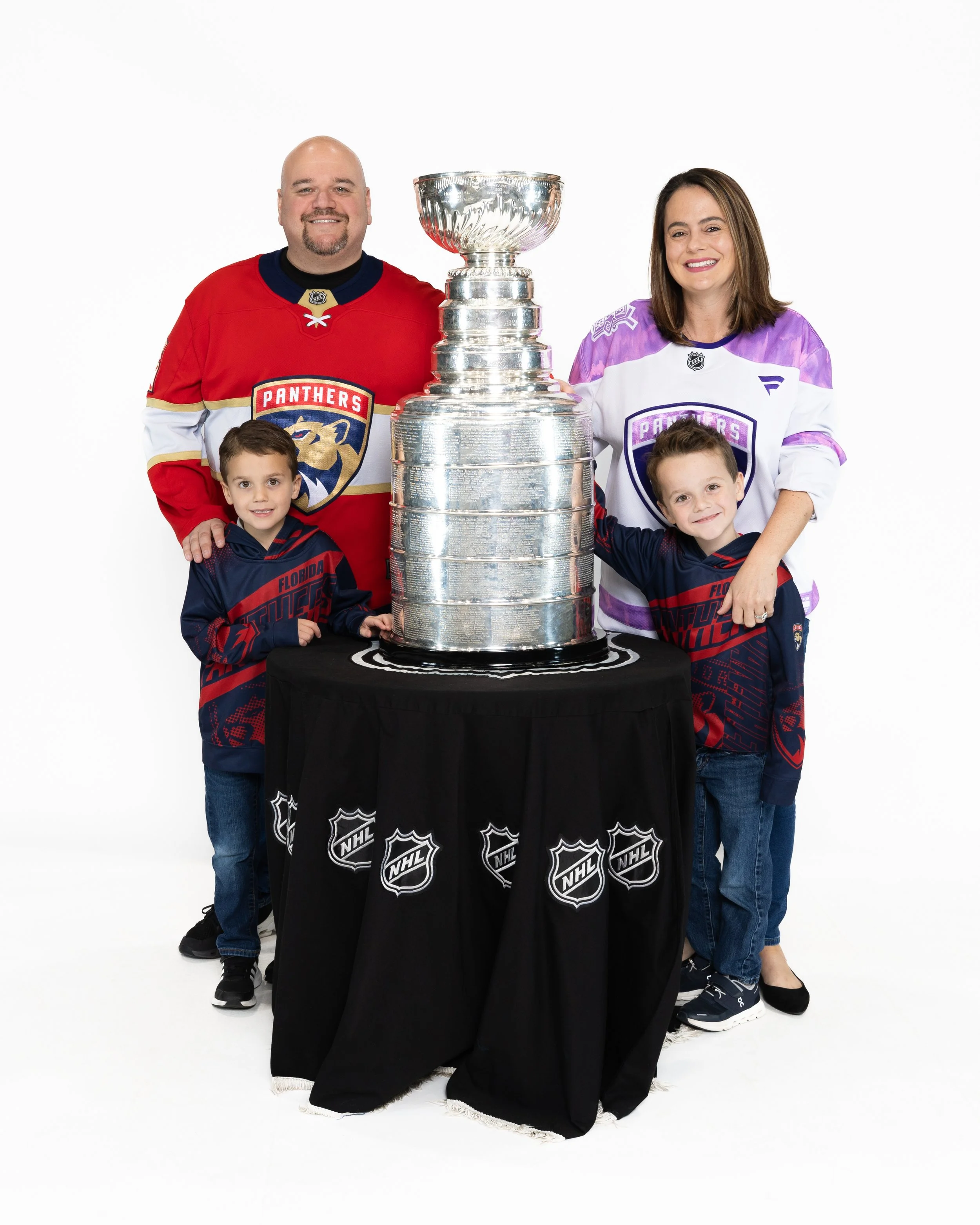 A family of four standing around the Stanley Cup, wearing hockey jerseys of the Florida Panthers, with a white background.