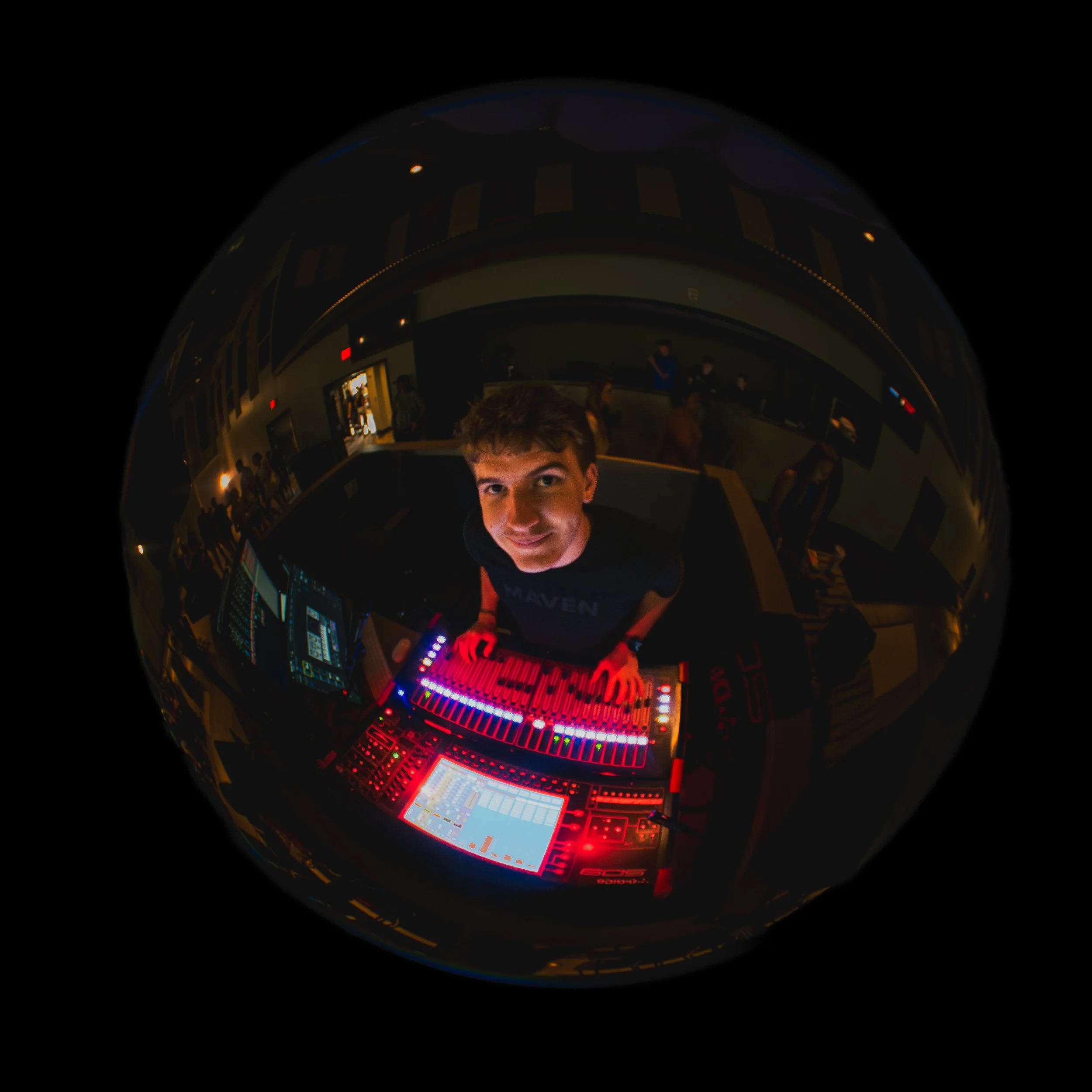 A young man with short brown hair, smiling, standing behind a DJ console with colorful lights in a dark room filled with people.