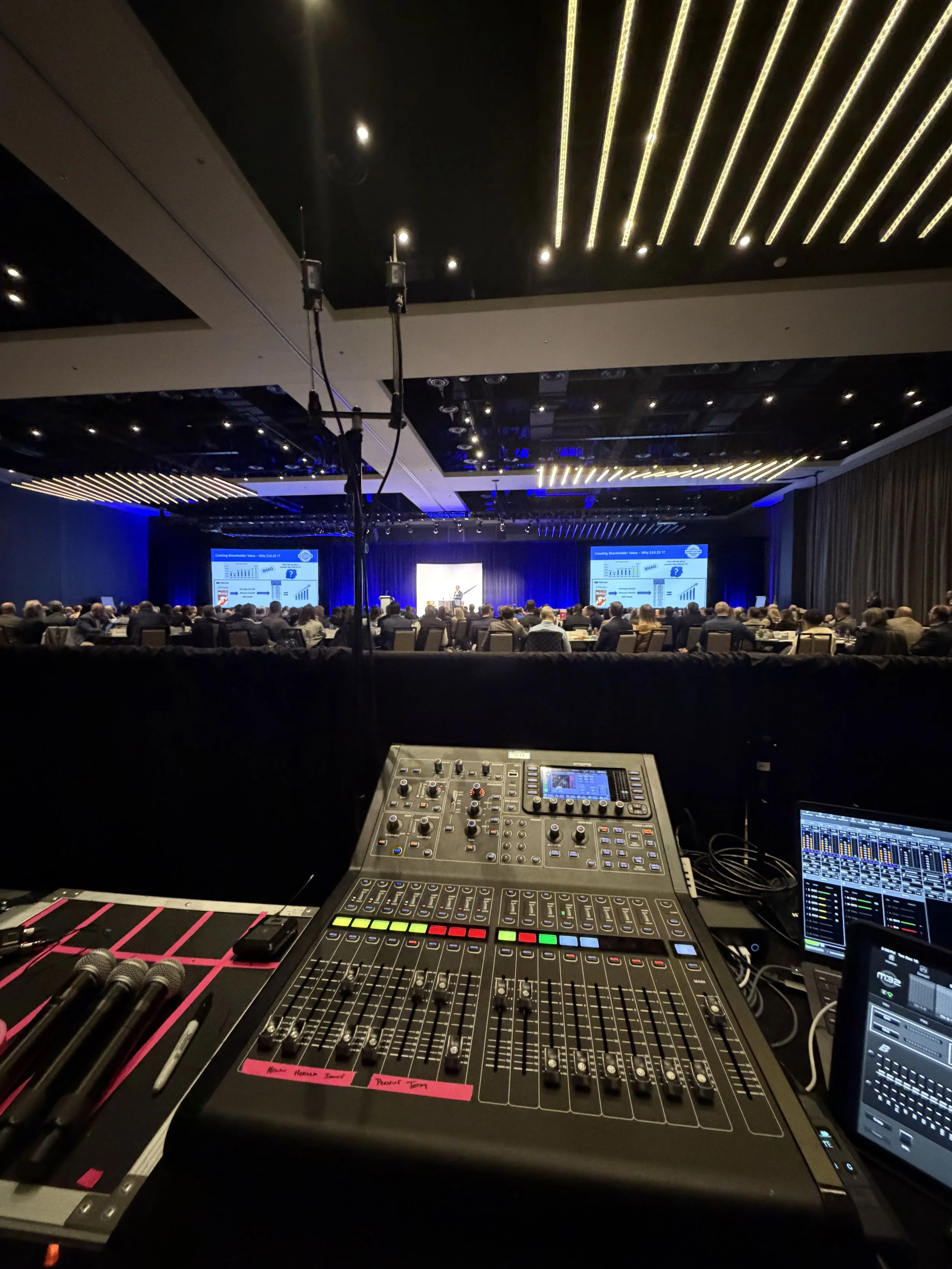 Behind-the-scenes view of a conference or presentation with a large audience, stage, and projection screens, taken from a sound engineer's perspective, showing mixing console and audio equipment.