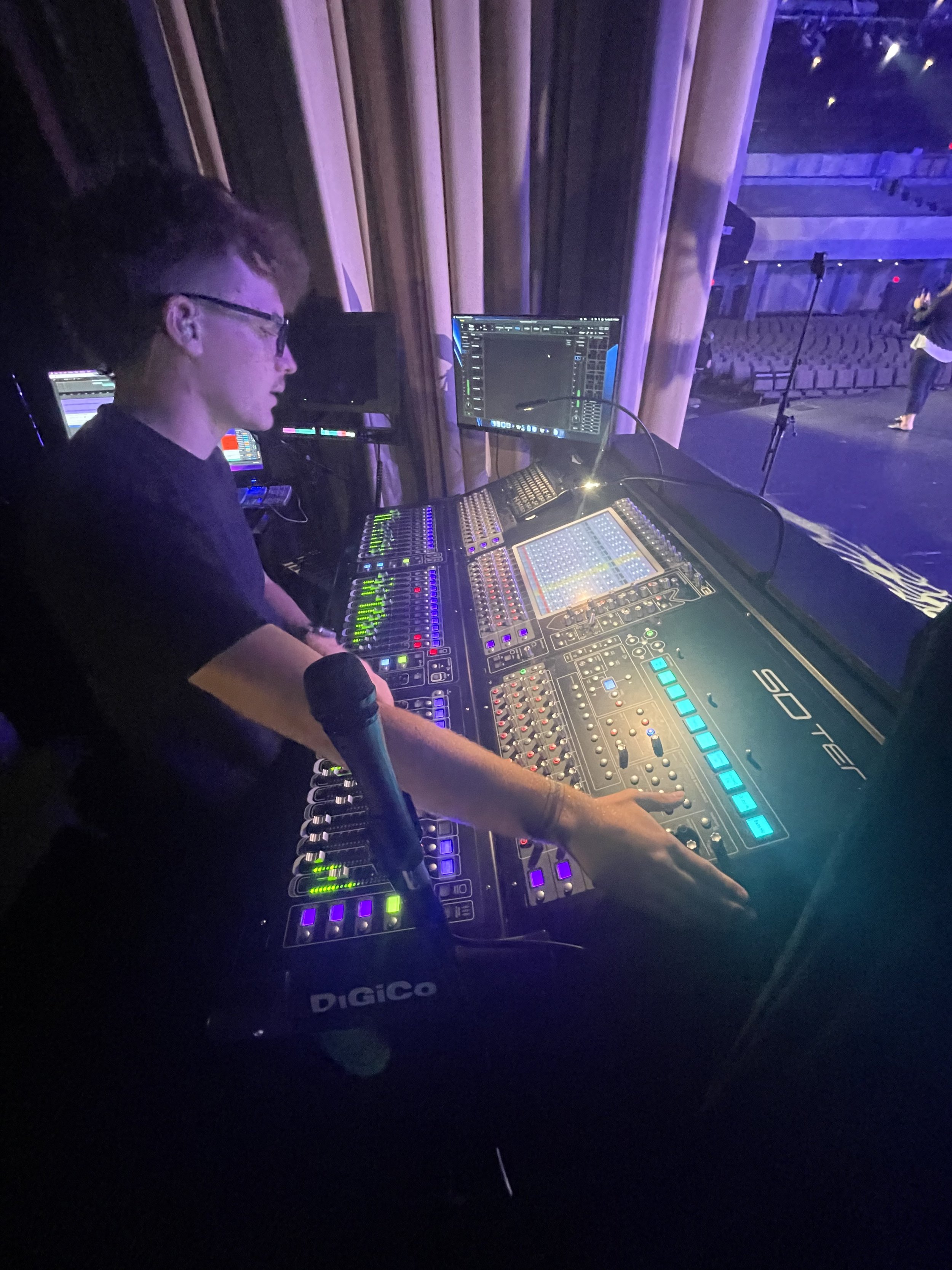 A sound engineer working at a mixing console on a stage, with stage lights and empty seats in the background.