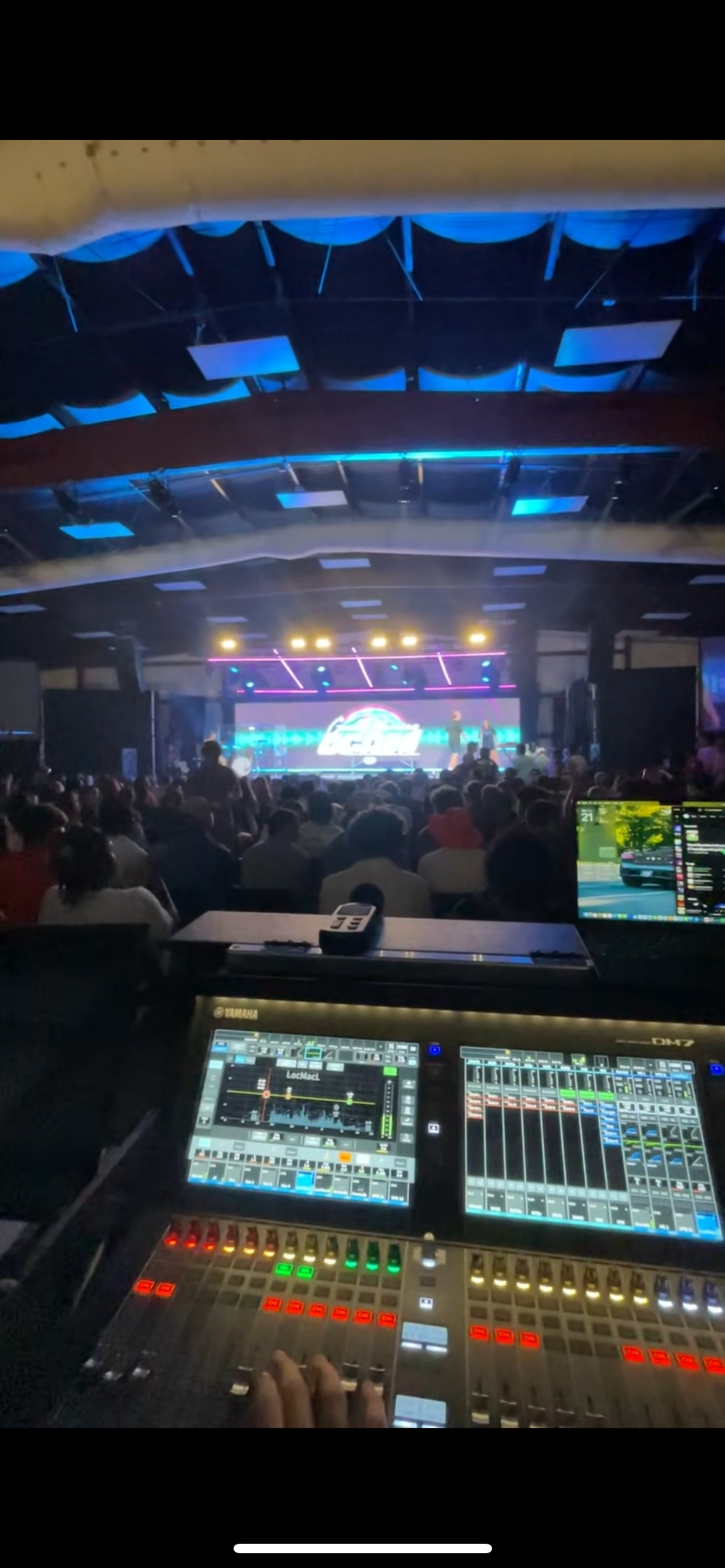 View of a large indoor concert with a crowd in front of a illuminated stage featuring digital screens and colorful lights, with a control panel and DJ equipment in the foreground.