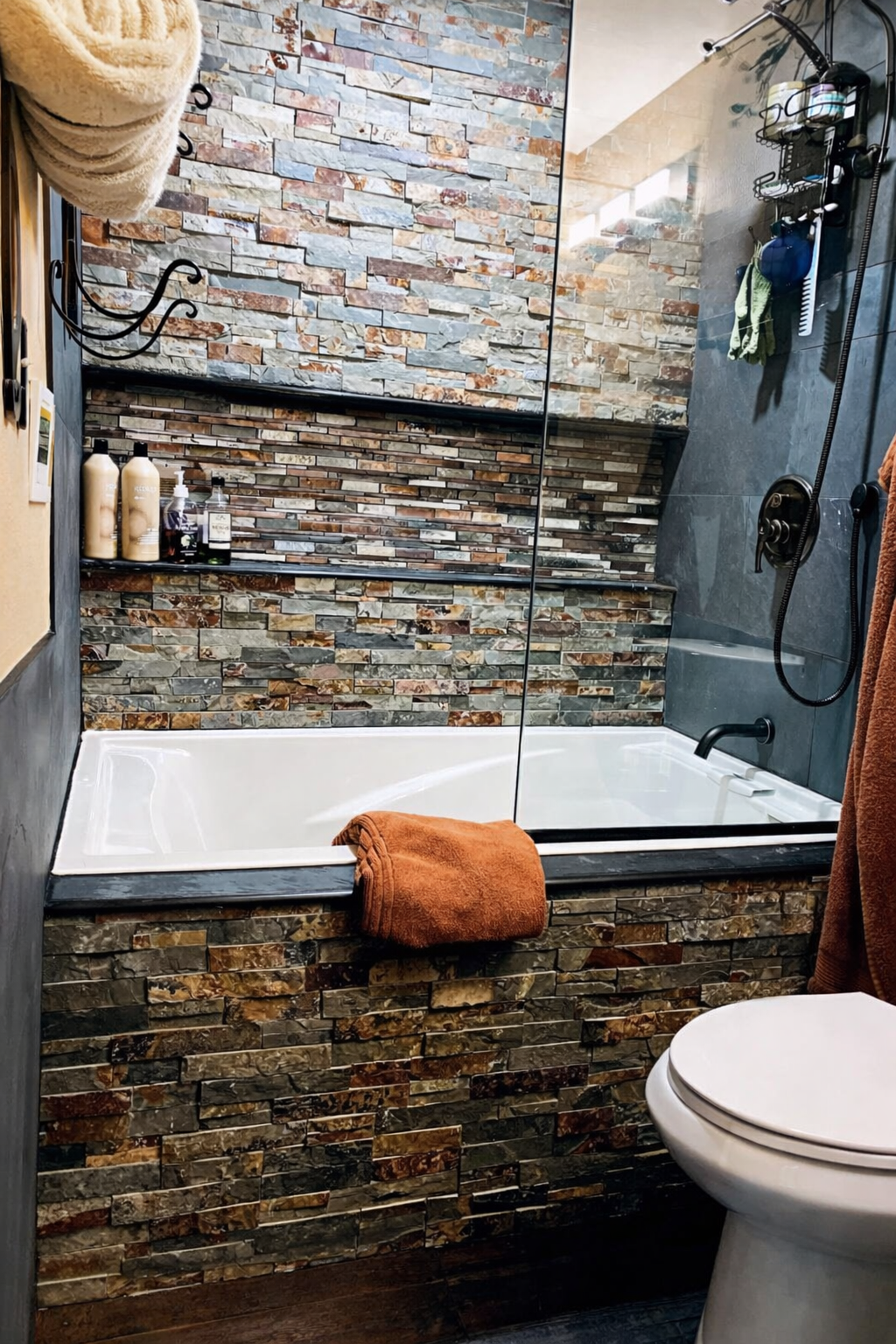 A modern bathroom featuring a bathtub with a partial stone tile surround, a glass shower wall, and a black showerhead. There is a black soap shelf on the wall with toiletries, a towel hanging on the side, and a rolled orange towel on the edge of the bathtub. The wall behind the bathtub is decorated with multicolored stone tiles.