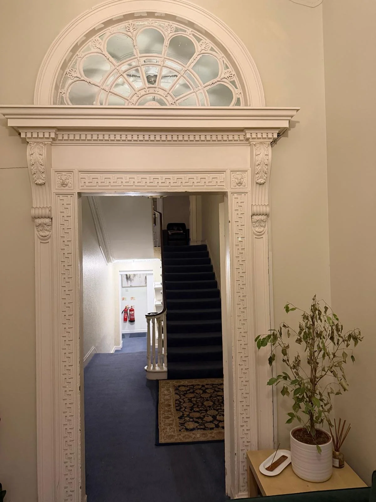 An ornate white doorframe with decorative moldings and a semi-circular window above it. Inside, a staircase with dark carpet leads upward, with a small rug at the entrance. To the right, a potted plant and a small table with a incense holder are visible.
